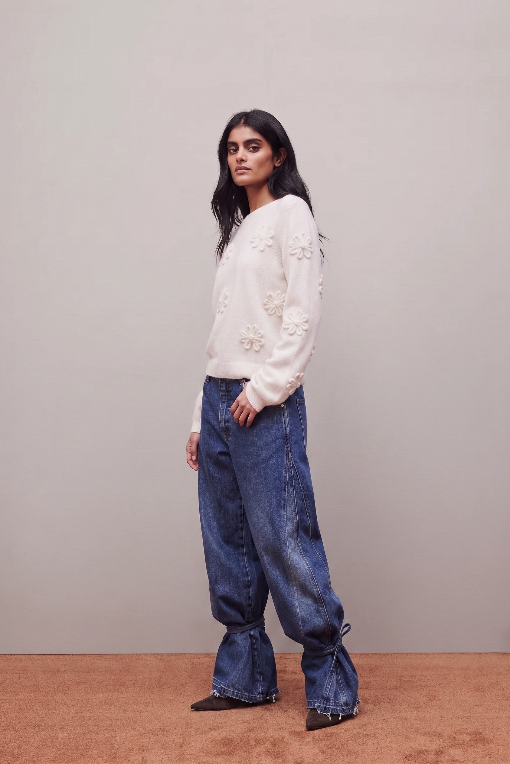 A woman modeling a cream-colored long-sleeve knit sweater with 3D floral embroidery and wide-leg blue jeans with gathered flared cuffs, standing against a neutral studio backdrop.