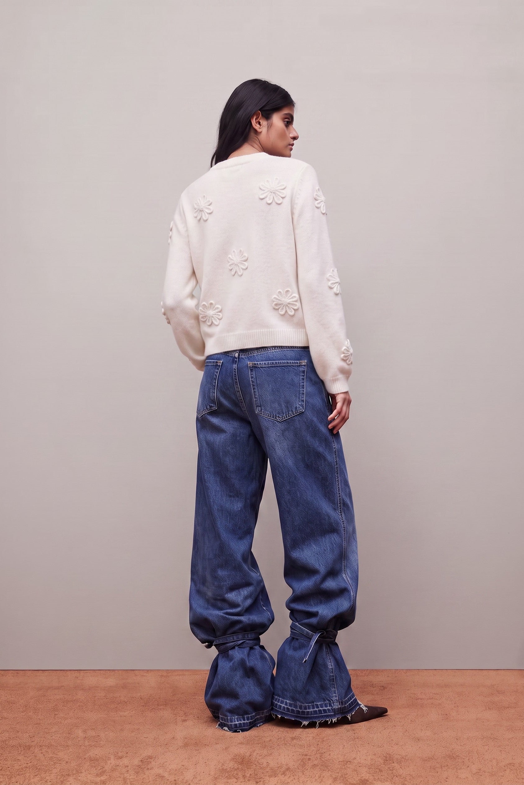 A model is shown from the back wearing a cream-colored knit sweater with raised floral embroidery and wide-leg blue jeans with frayed hems, standing against a neutral studio backdrop.