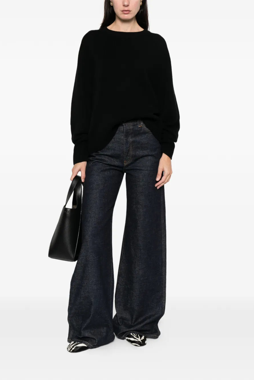 Woman wearing a black sweater and dark jeans holding a black handbag on a white background.