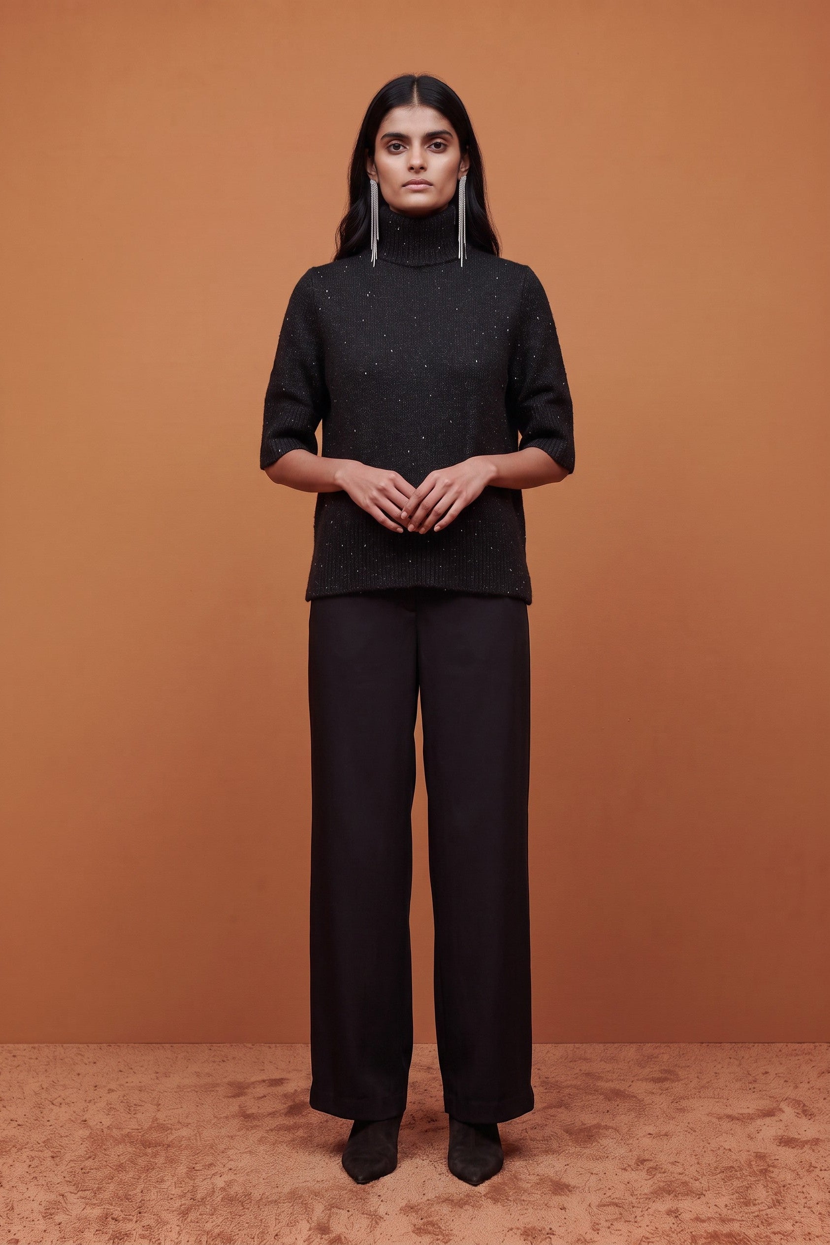 A model stands against a warm brown studio backdrop wearing a black turtleneck top with subtle sparkly texture, black wide-leg trousers, and long silver earrings.