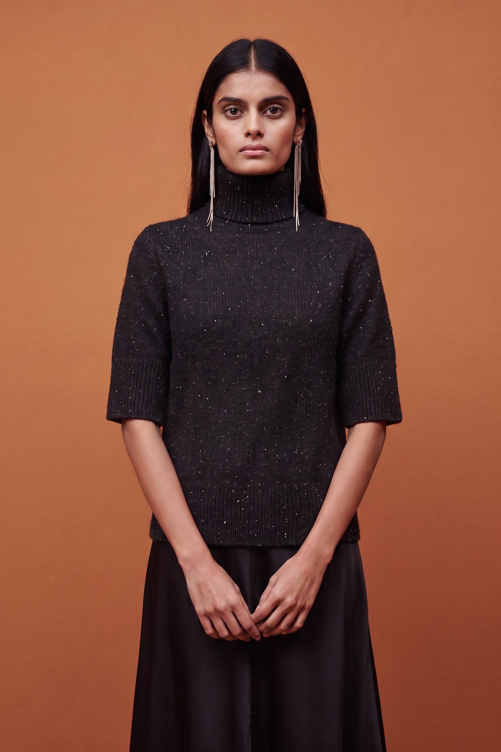 A model stands against a solid orange-brown studio background wearing a dark charcoal gray short-sleeve turtleneck sweater with subtle sparkly flecks and a black skirt.