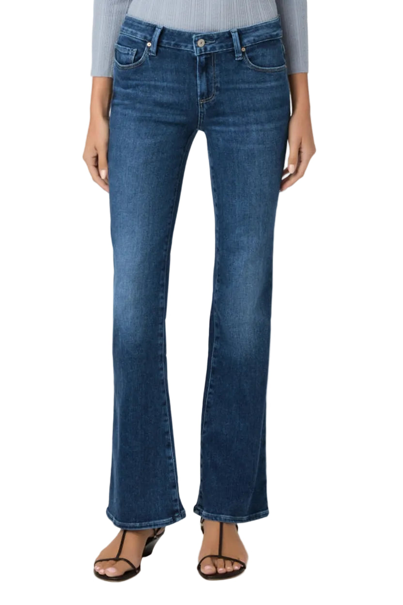 A model wearing a pair of medium-wash blue flare jeans, styled with a light gray ribbed top and brown strappy sandals, against a plain light gray studio background.