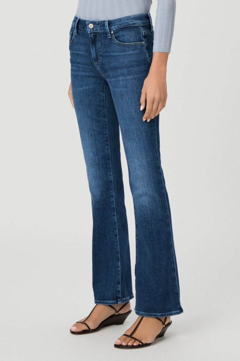 A model wearing a pair of medium-wash blue flare jeans, styled with a light blue ribbed top and black wedge sandals, photographed against a clean white studio background.