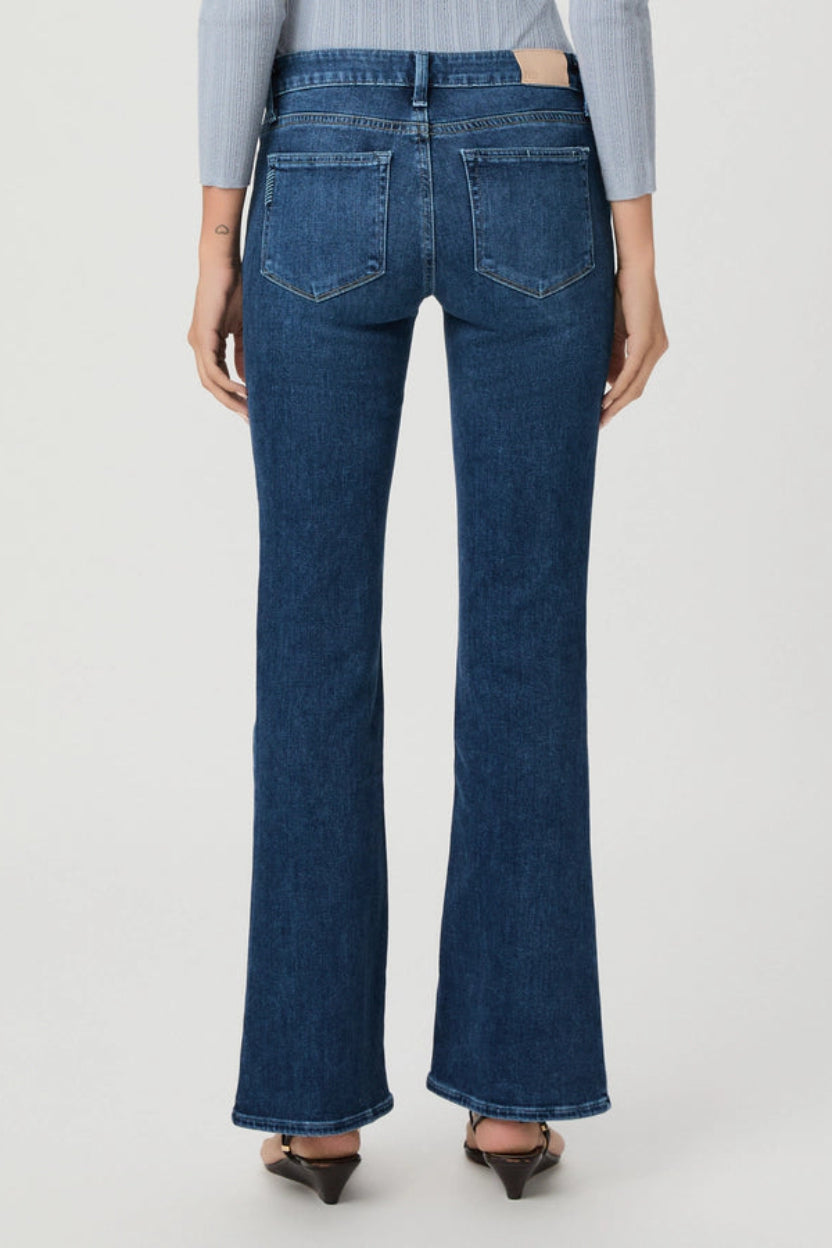 Back view of a model wearing dark blue flare jeans with a light blue striped top and brown pointed-toe heels, set against a plain light gray studio background.