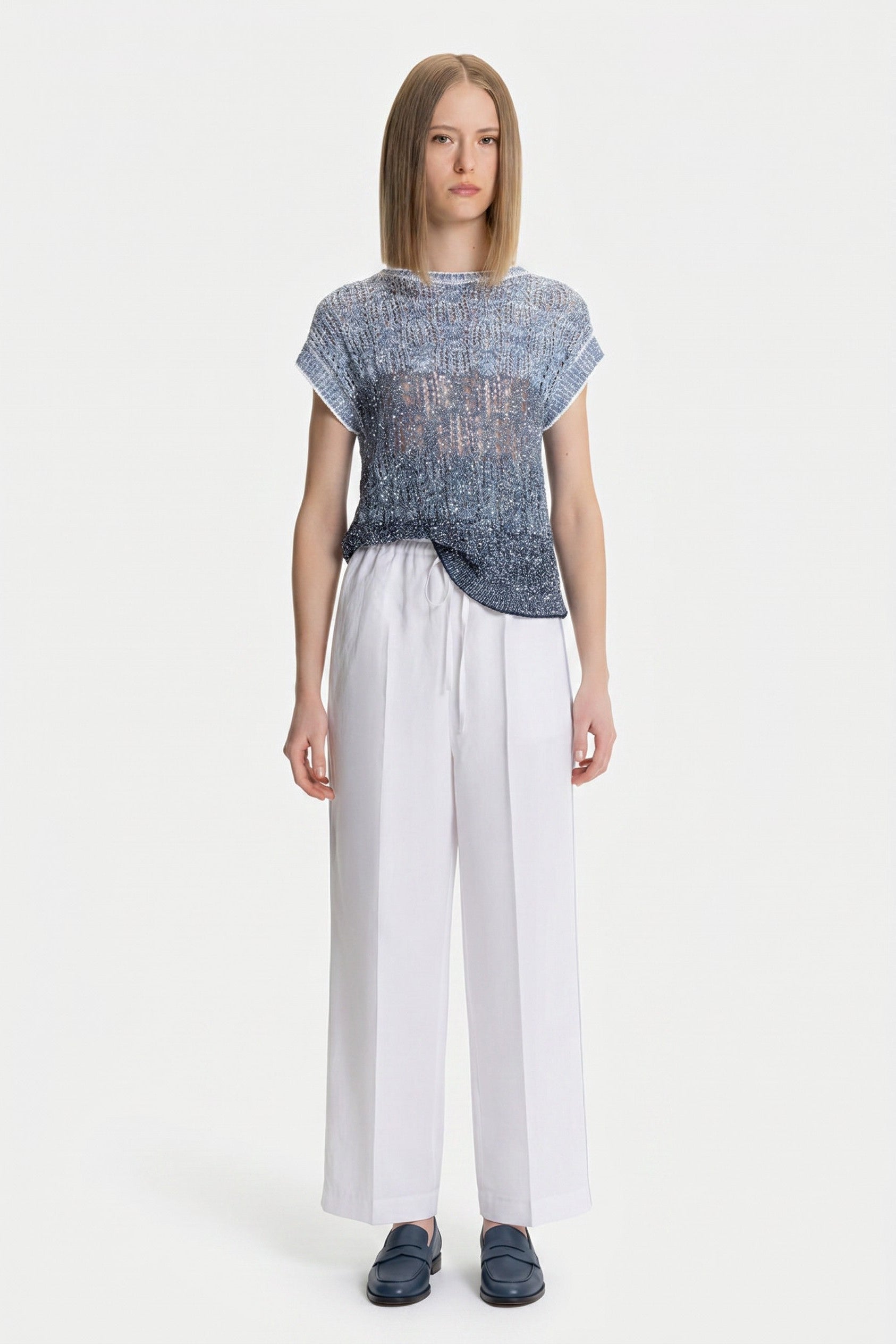 Woman wearing a blue knitted sparkle top and white pants on a white background.