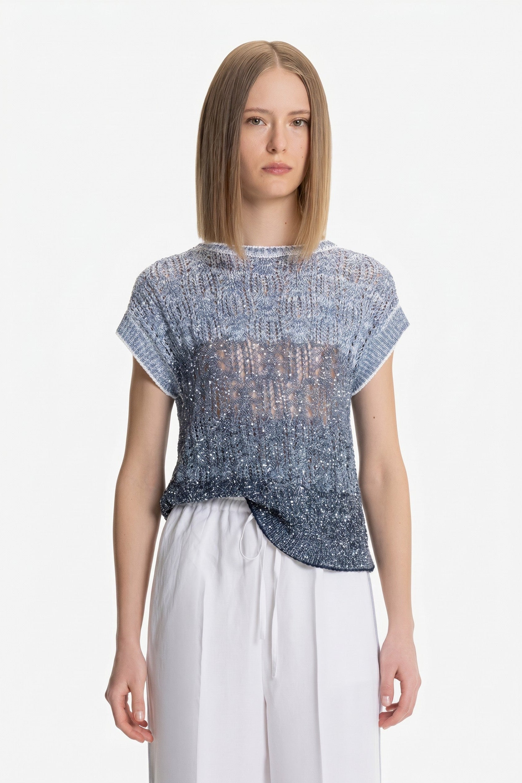 Woman wearing a blue patterned top and white pants on a white background.