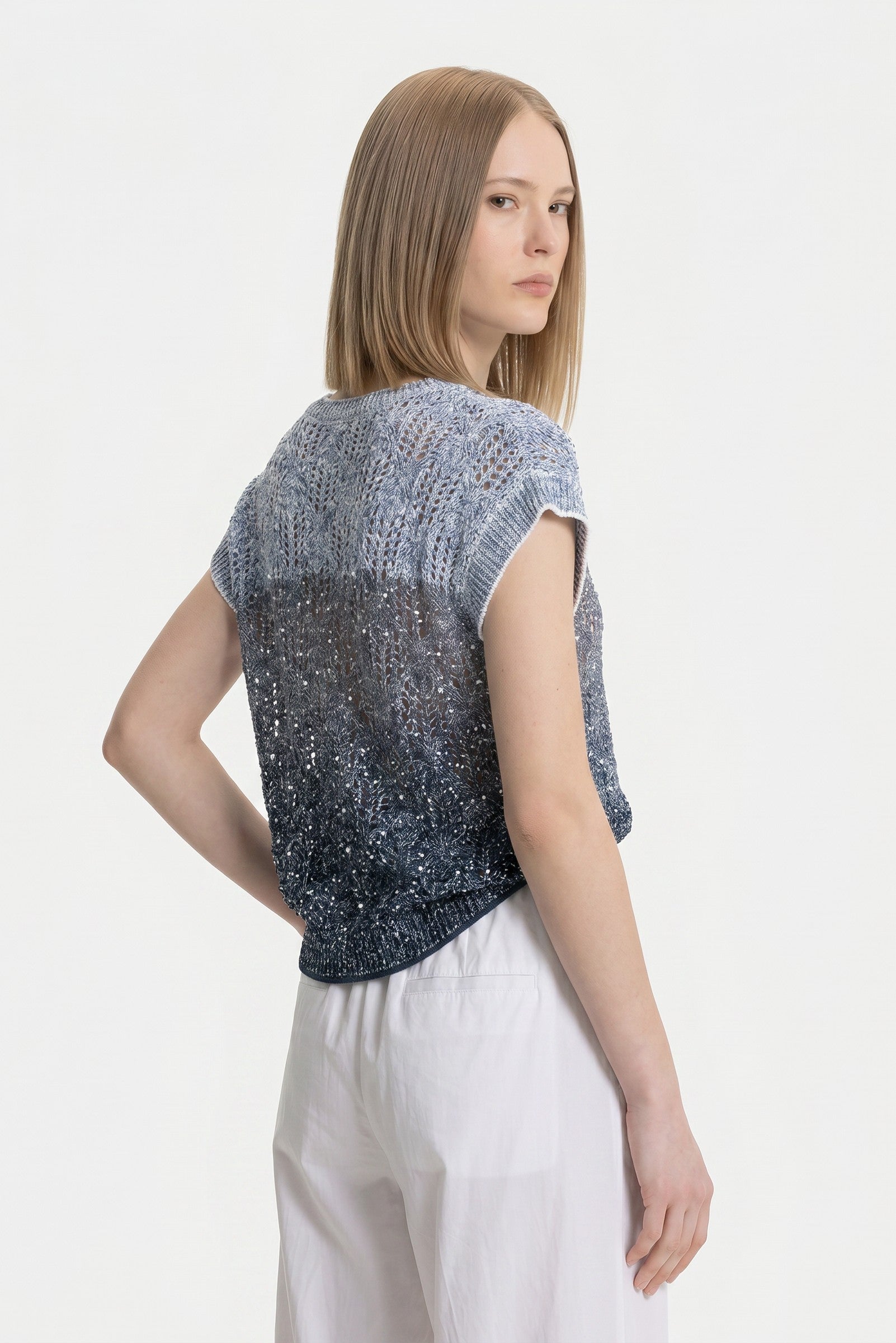 The back of a woman wearing a blue sequin patterned top with white pants on a white background.