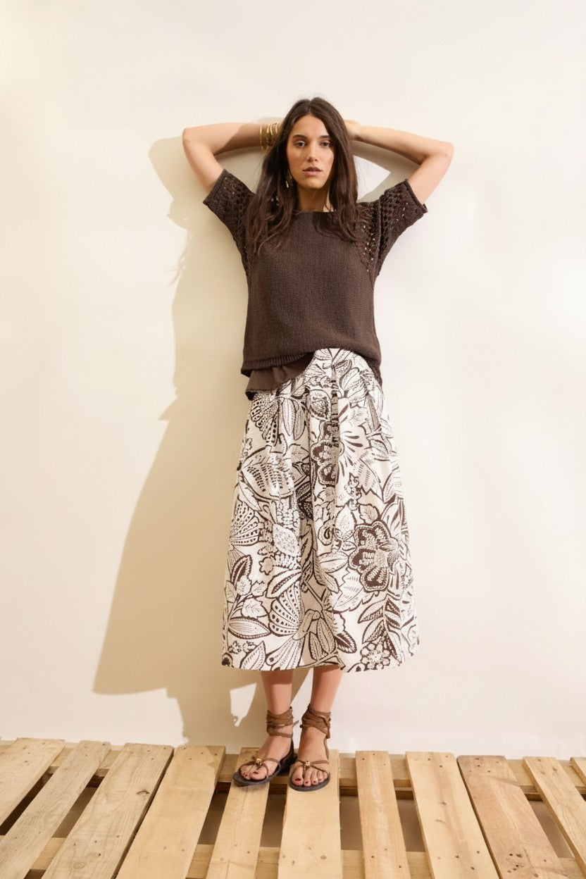 Woman wearing a brown top and a white and brown pattern midi skirt standing on a wooden platform with a beige wall background.