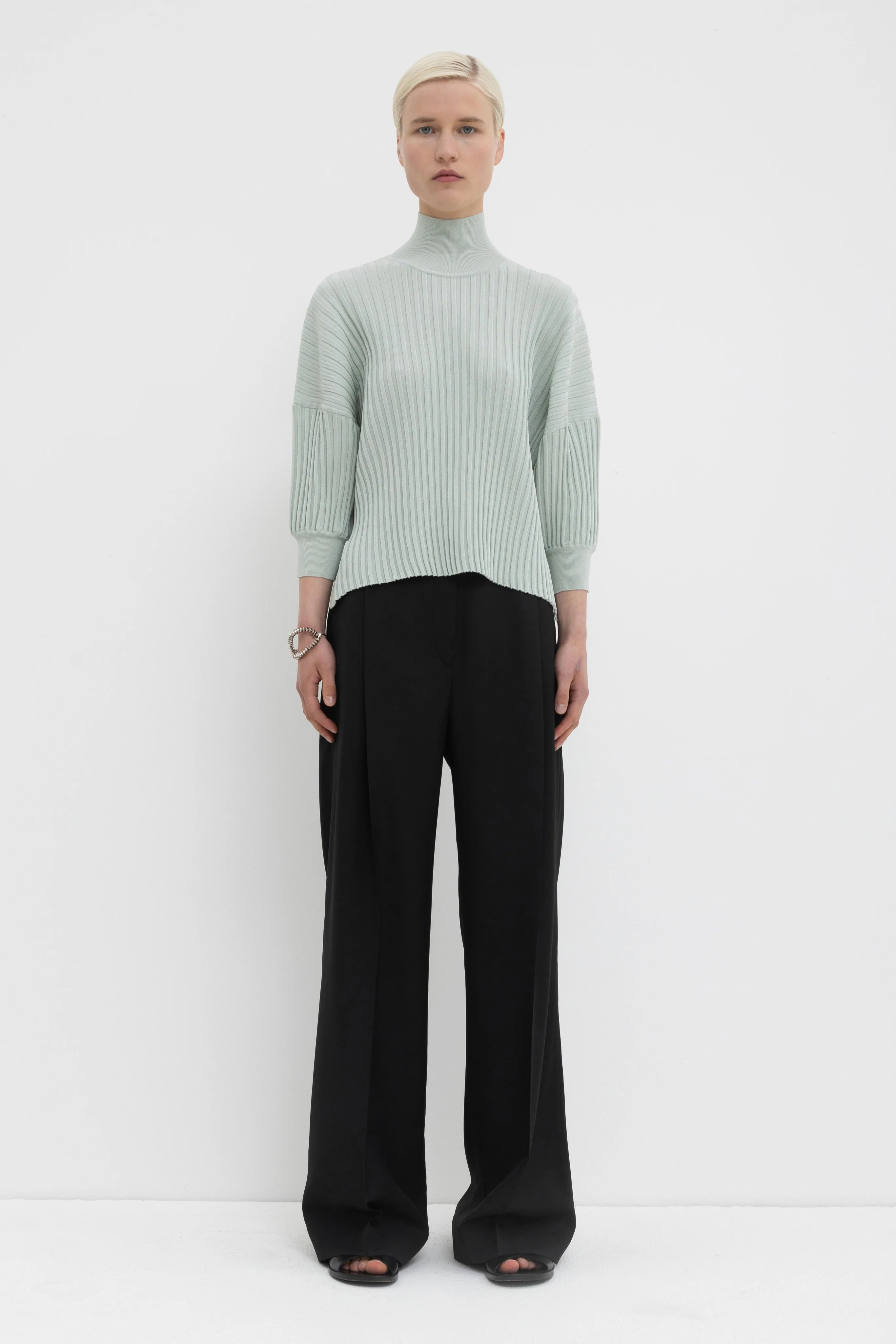 Person wearing a light green ribbed sweater and black pants on a white background