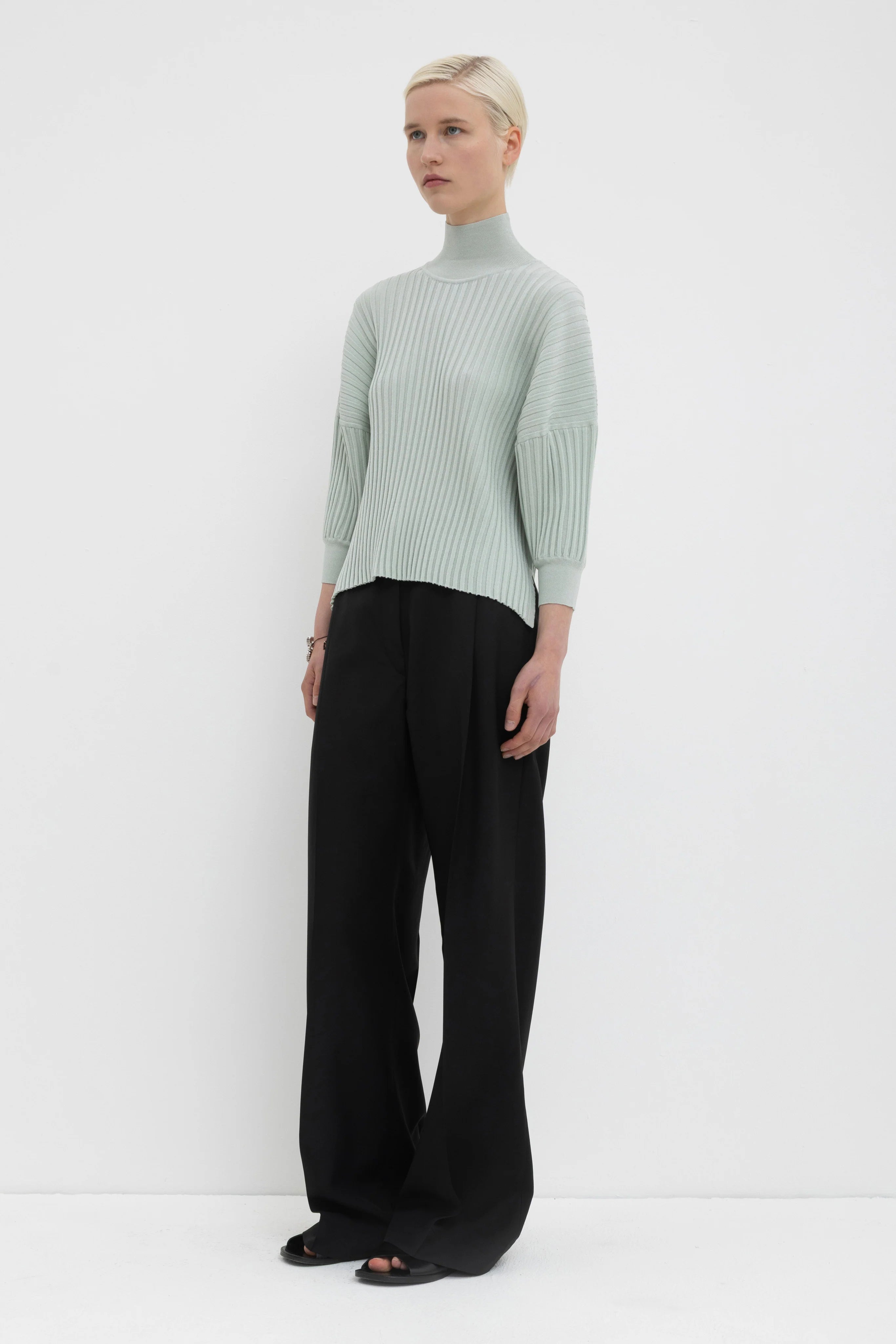 Person wearing a light green ribbed sweater and black pants on a white background