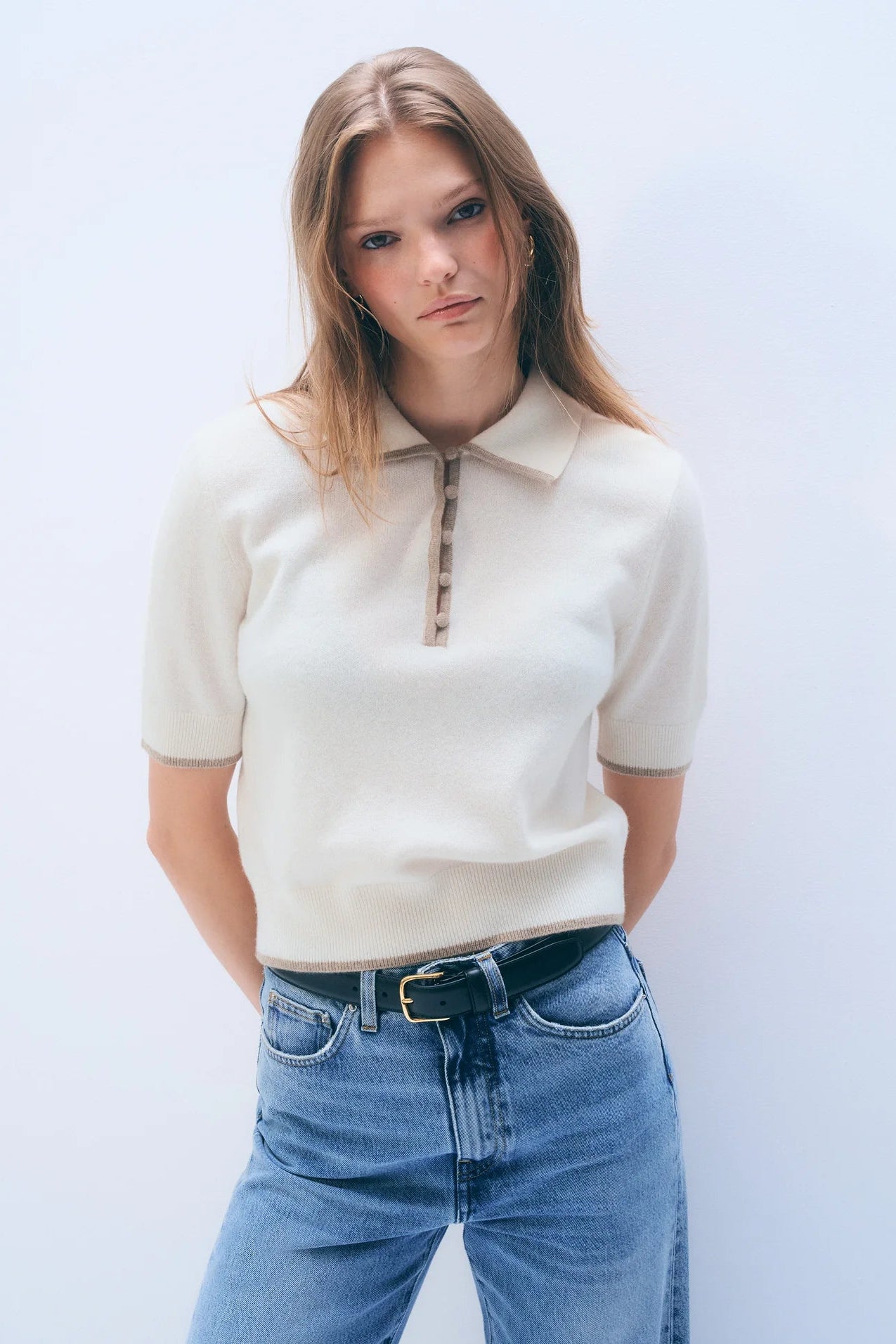 Woman wearing a cream polo shirt and blue jeans against a white background.