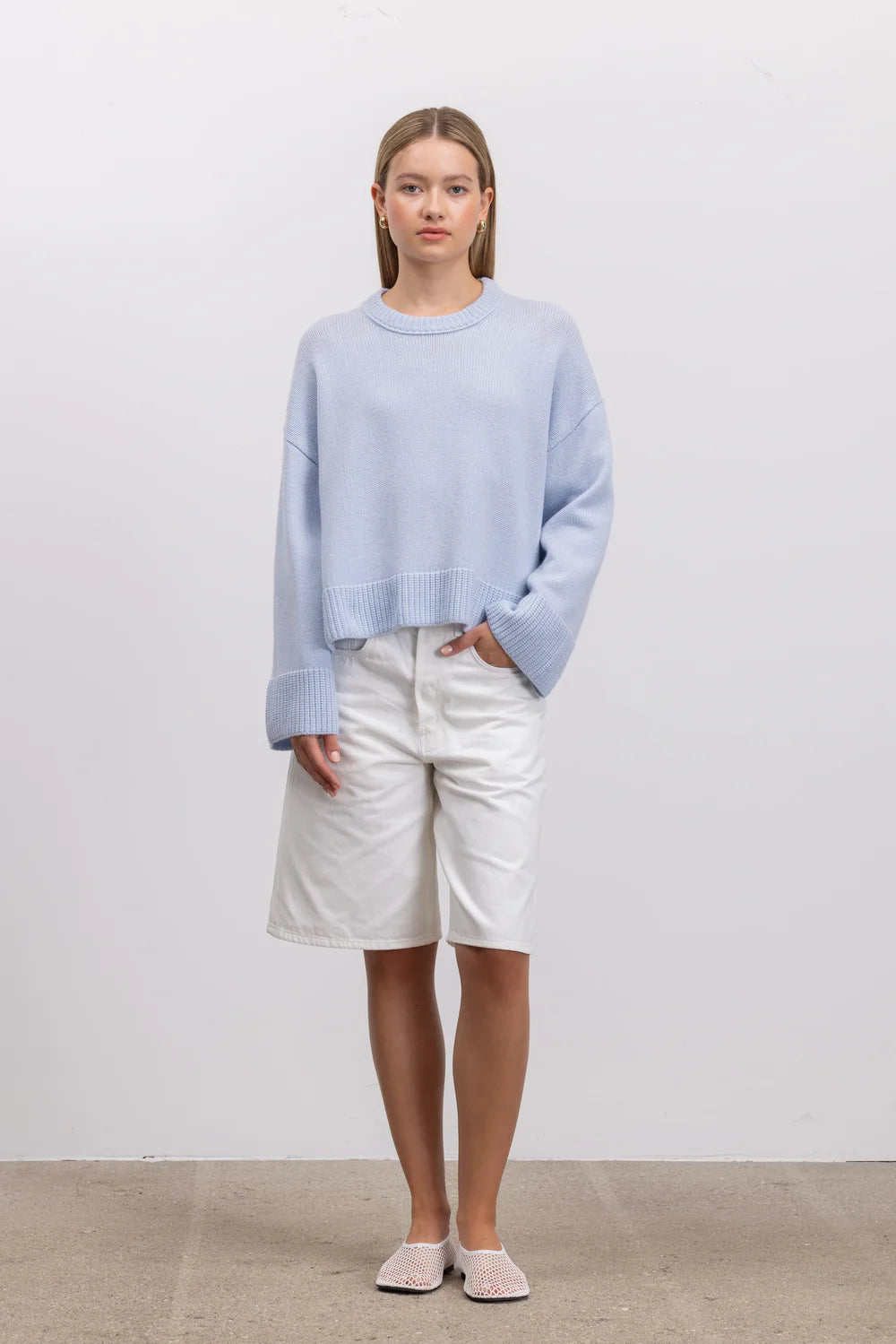 A woman stands against a plain light gray wall wearing a light blue cropped crewneck sweater, white shorts, and pale pink slip-on shoes.