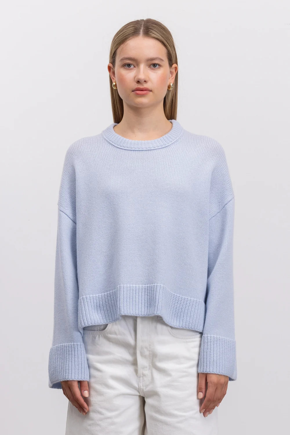 A woman wearing a light blue cropped knit sweater with ribbed cuffs and hem, paired with white pants, standing against a plain light gray studio background.