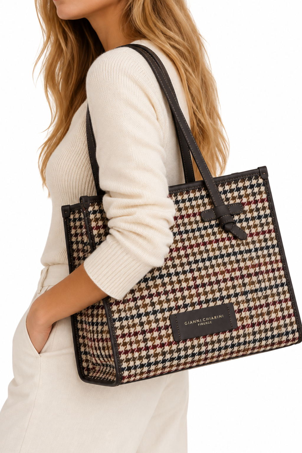 A woman wearing a cream ribbed sweater and white dress carries a large houndstooth tote bag with black leather trim and shoulder straps. The bag features a black leather brand patch.