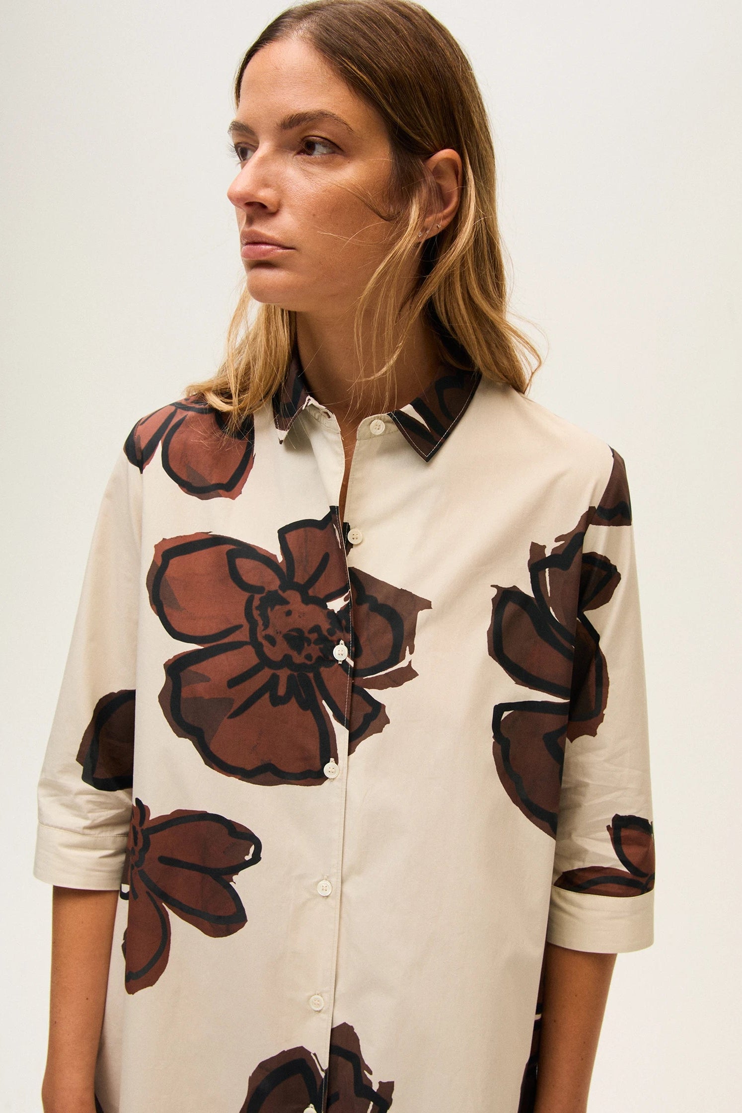 Close-up of a woman wearing a cream dress with brown floral patterns on a plain background.