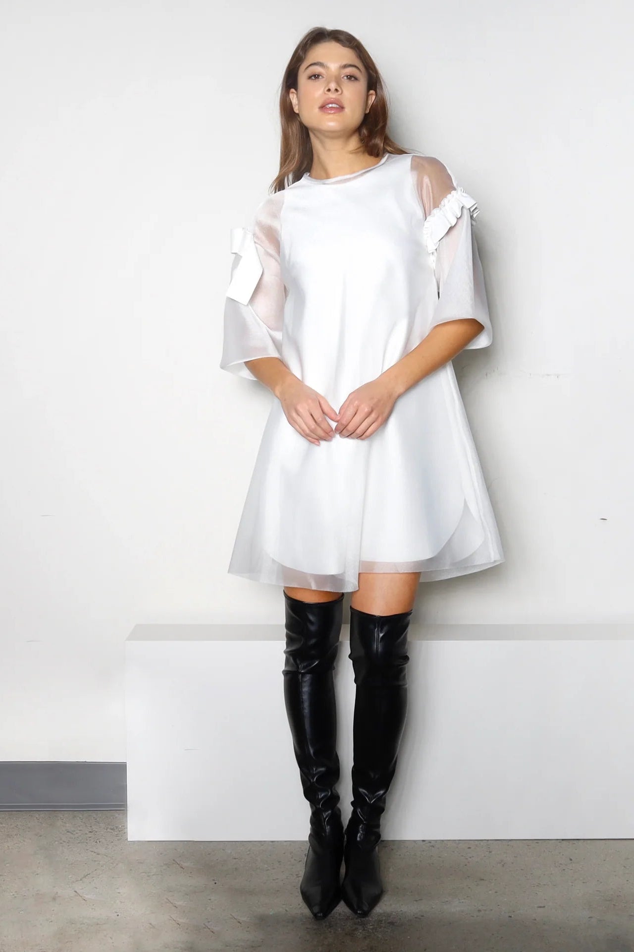 Woman wearing a white dress with sheer shoulders and black knee-high boots against a plain background.