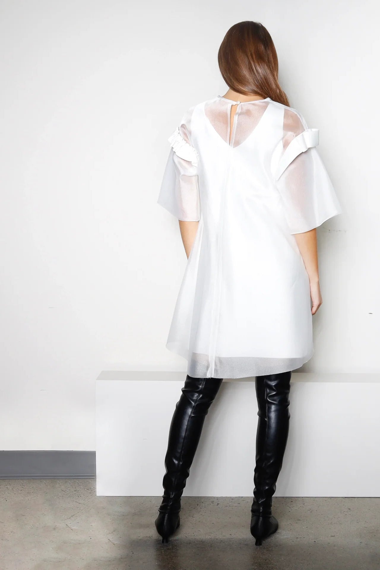 The back of a woman wearing a white dress with sheer sleeves and long black boots against a plain wall.