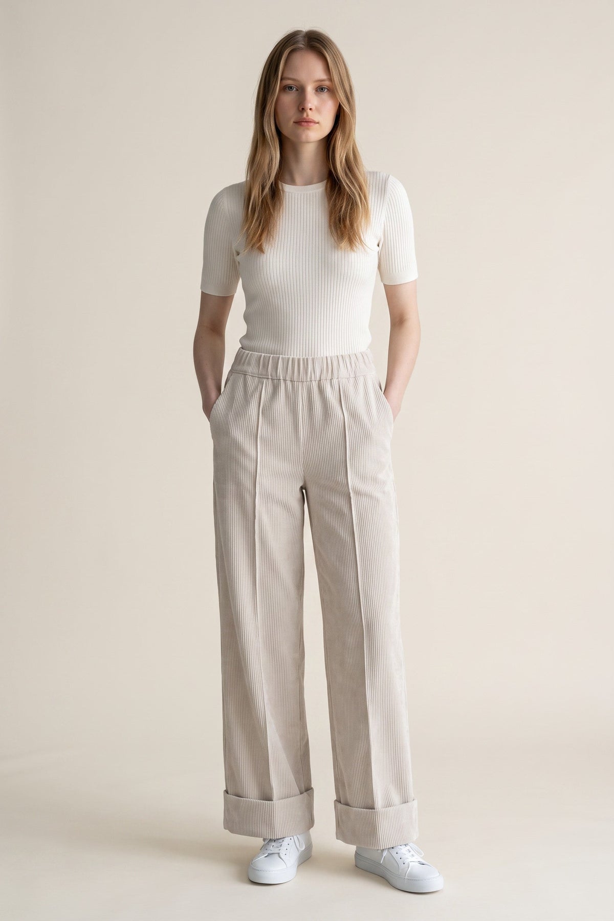 A woman modeling a white short-sleeve ribbed knit top and light beige wide-leg trousers with rolled cuffs, paired with white sneakers, against a solid light beige studio background.