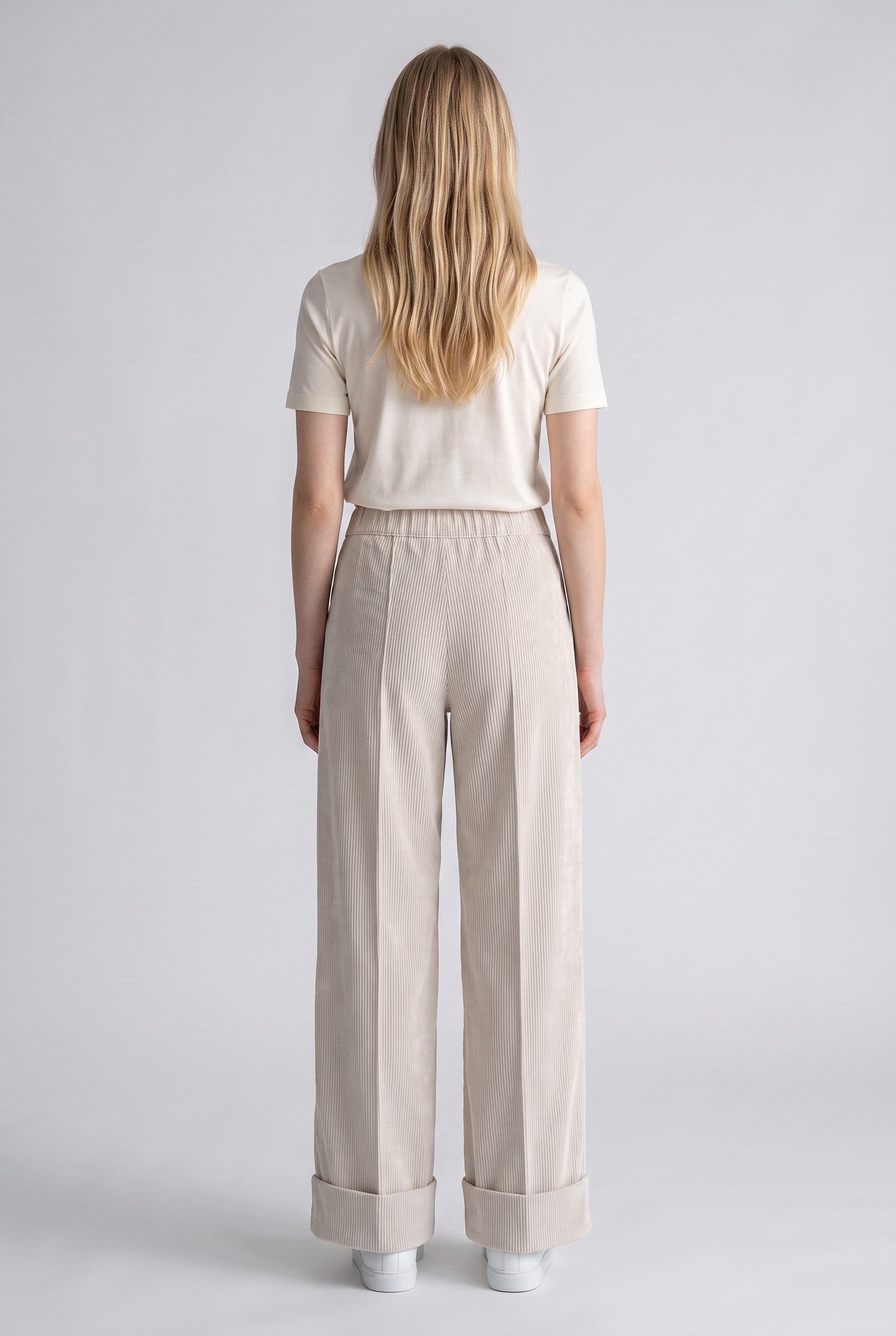 A woman viewed from the back wearing a cream short-sleeve top and light beige wide-leg trousers with rolled cuffs, standing against a light gray studio background.