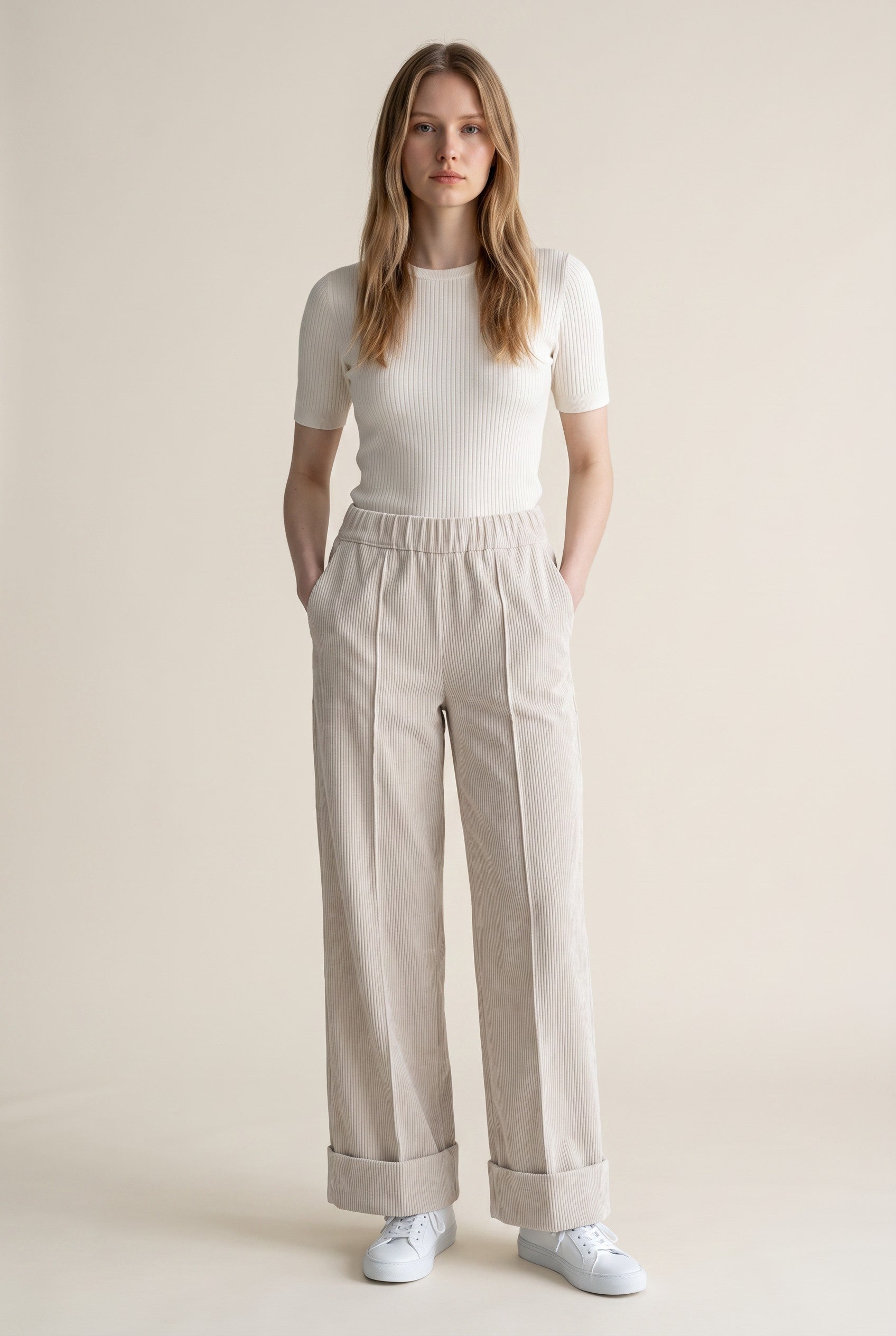 A woman modeling a white short-sleeve ribbed knit top and light beige wide-leg trousers with rolled cuffs, paired with white sneakers, against a solid light beige studio background.