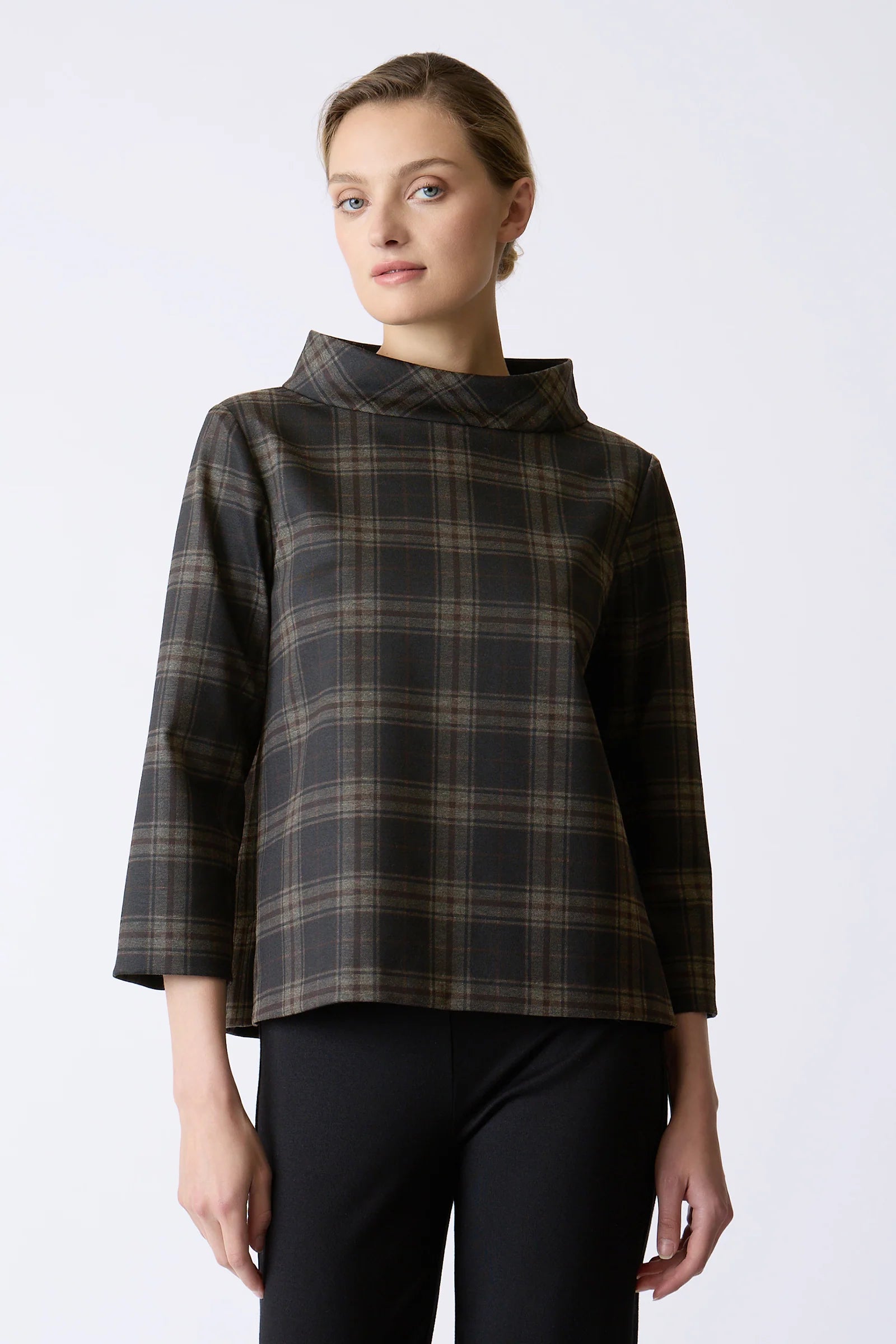 A woman modeling a dark brown and black plaid high-neck top with three-quarter sleeves, paired with black pants, against a plain light gray background.