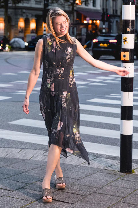 Woman in a black floral dress with platform sandals standing on a city street corner.