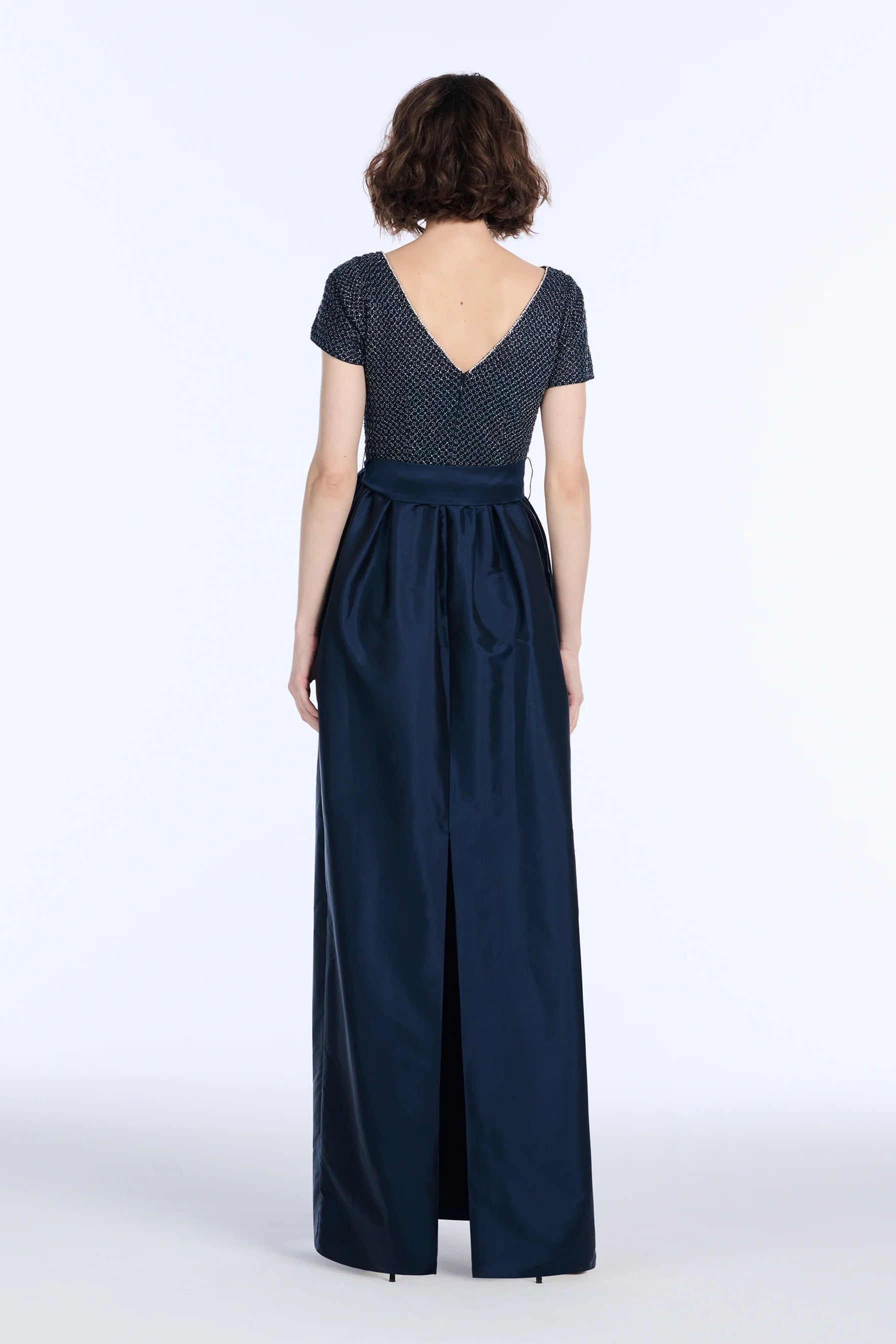 The back of a woman wearing a navy blue dress with a beaded top and plain skirt on a white background.