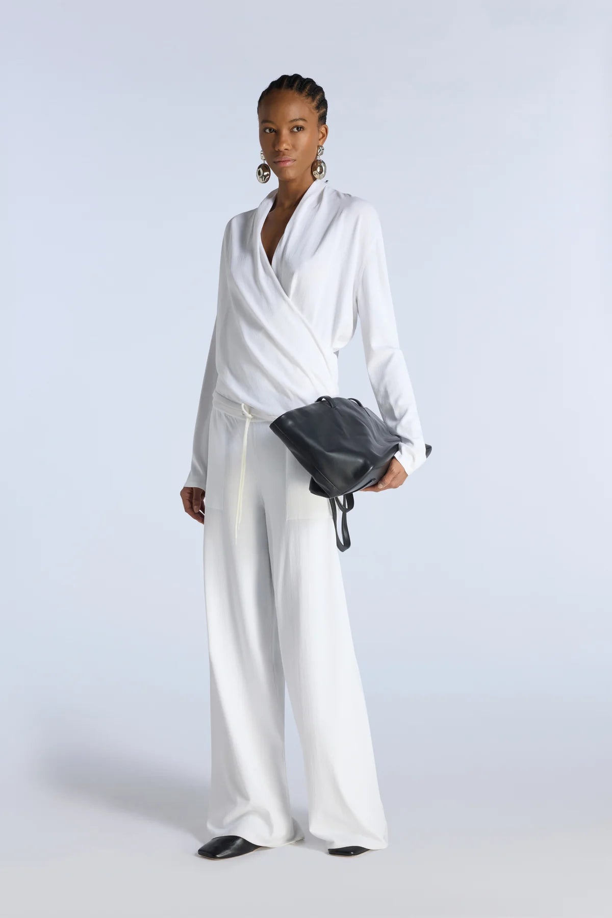 Woman in a white outfit holding a black bag against a light gray background