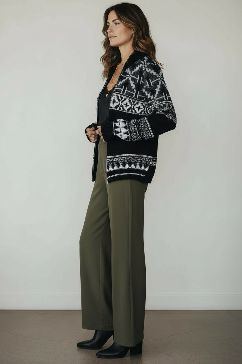 Woman wearing a black patterned zip up with green pants against a white background.