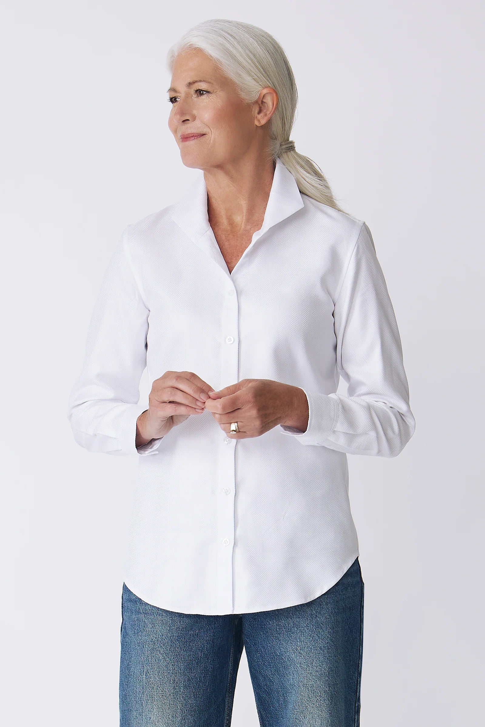 A woman with gray hair tied back wears a white long-sleeve button-up shirt and blue jeans, standing against a light gray studio background.