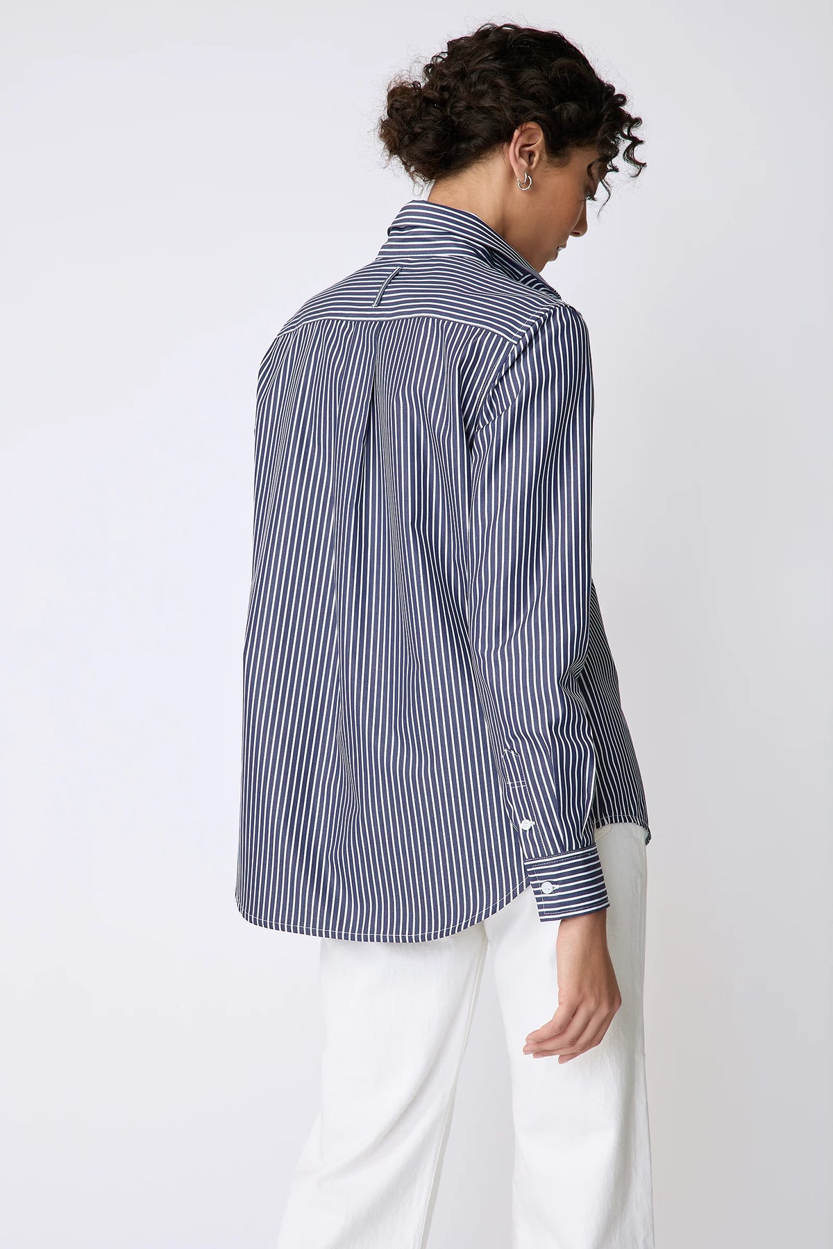A model shown from the back wearing a navy and white vertically striped button-down shirt and white trousers, photographed against a clean white studio background.