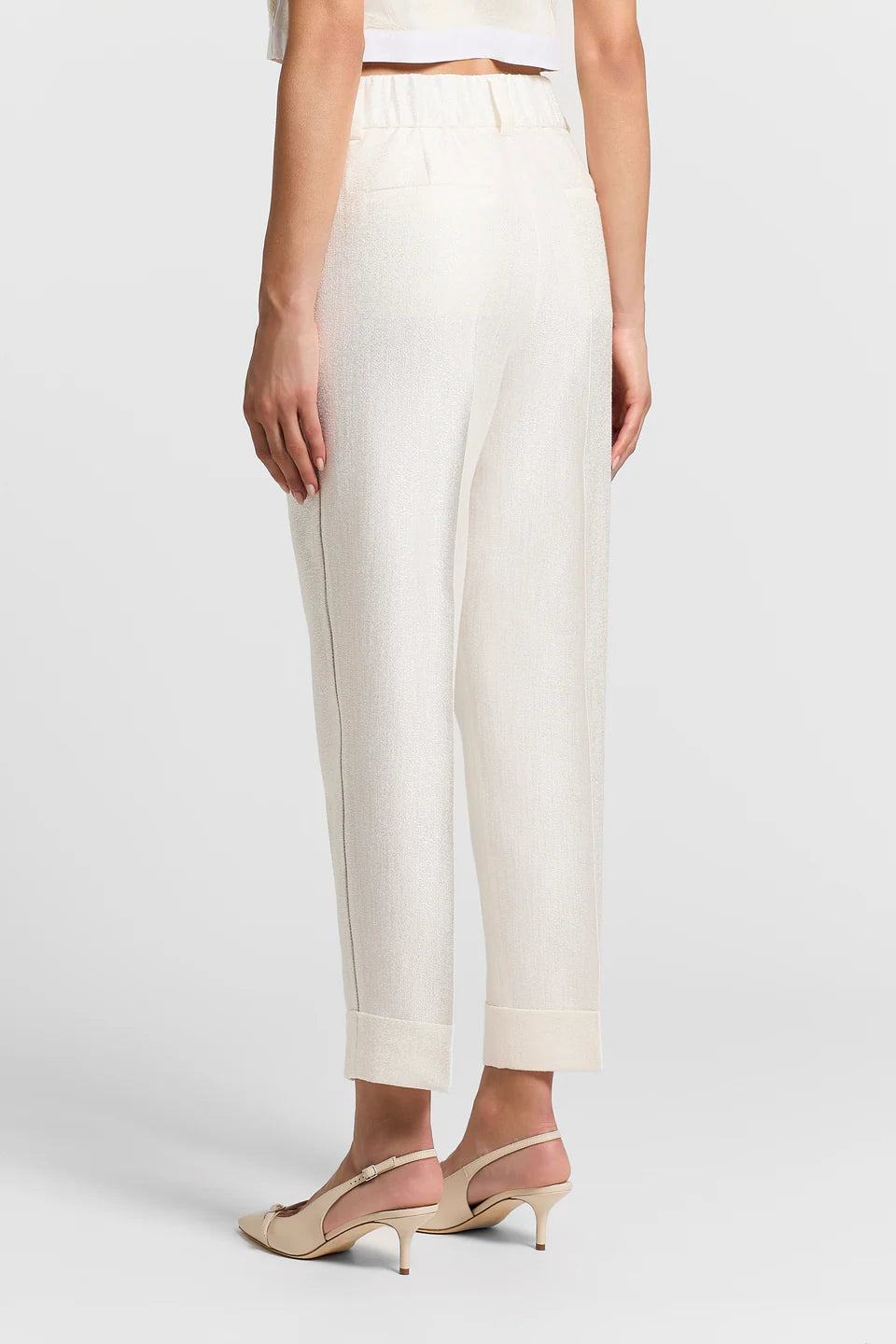 Back of a woman wearing white pants and cream heels on a plain background.