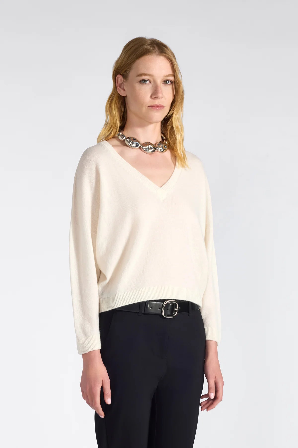 Woman wearing an off white sweater and black pants with a white background.