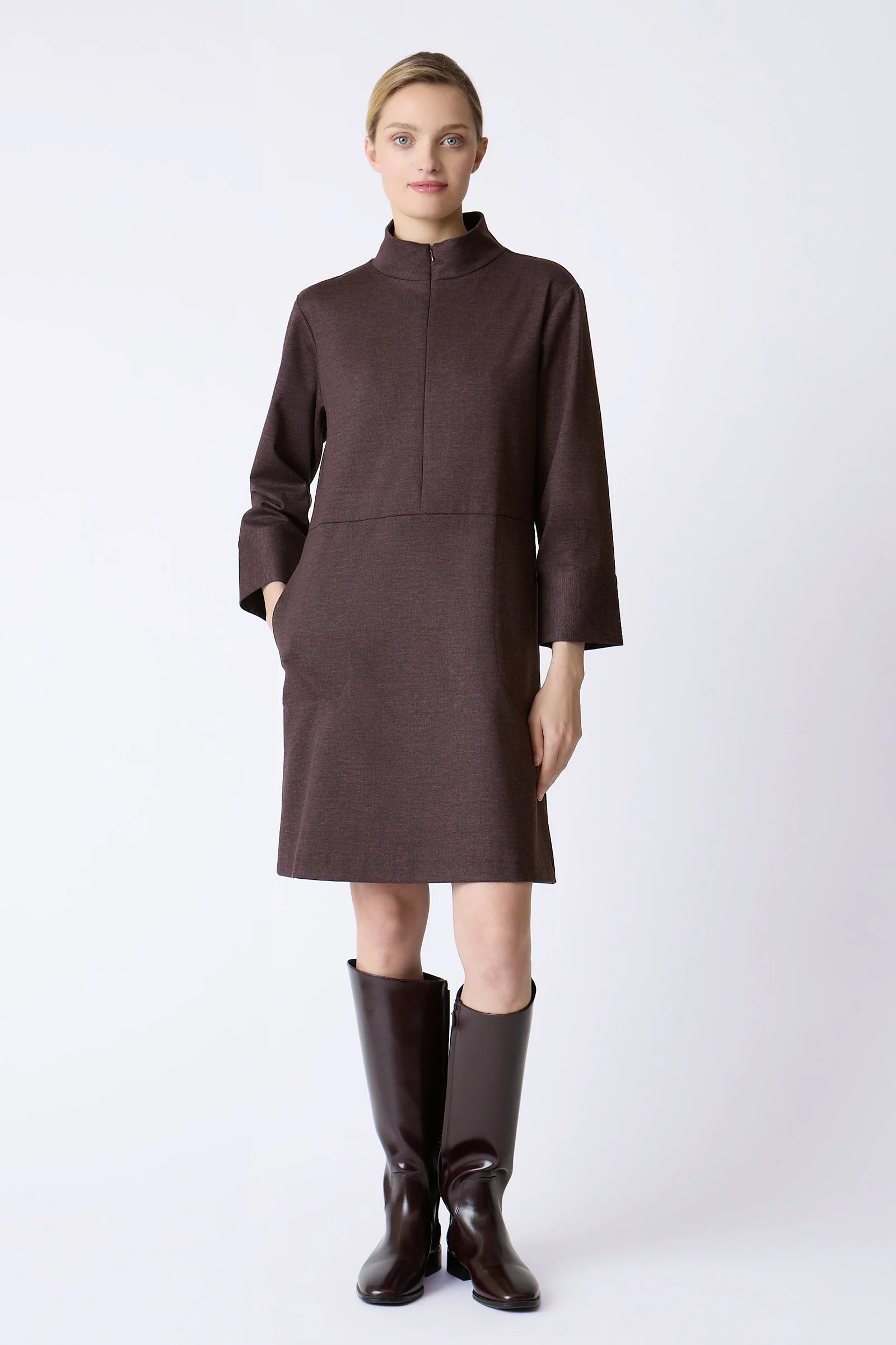A woman stands against a light gray studio background wearing a burgundy mock-neck long-sleeve dress with a straight hem and matching dark brown knee-high boots.