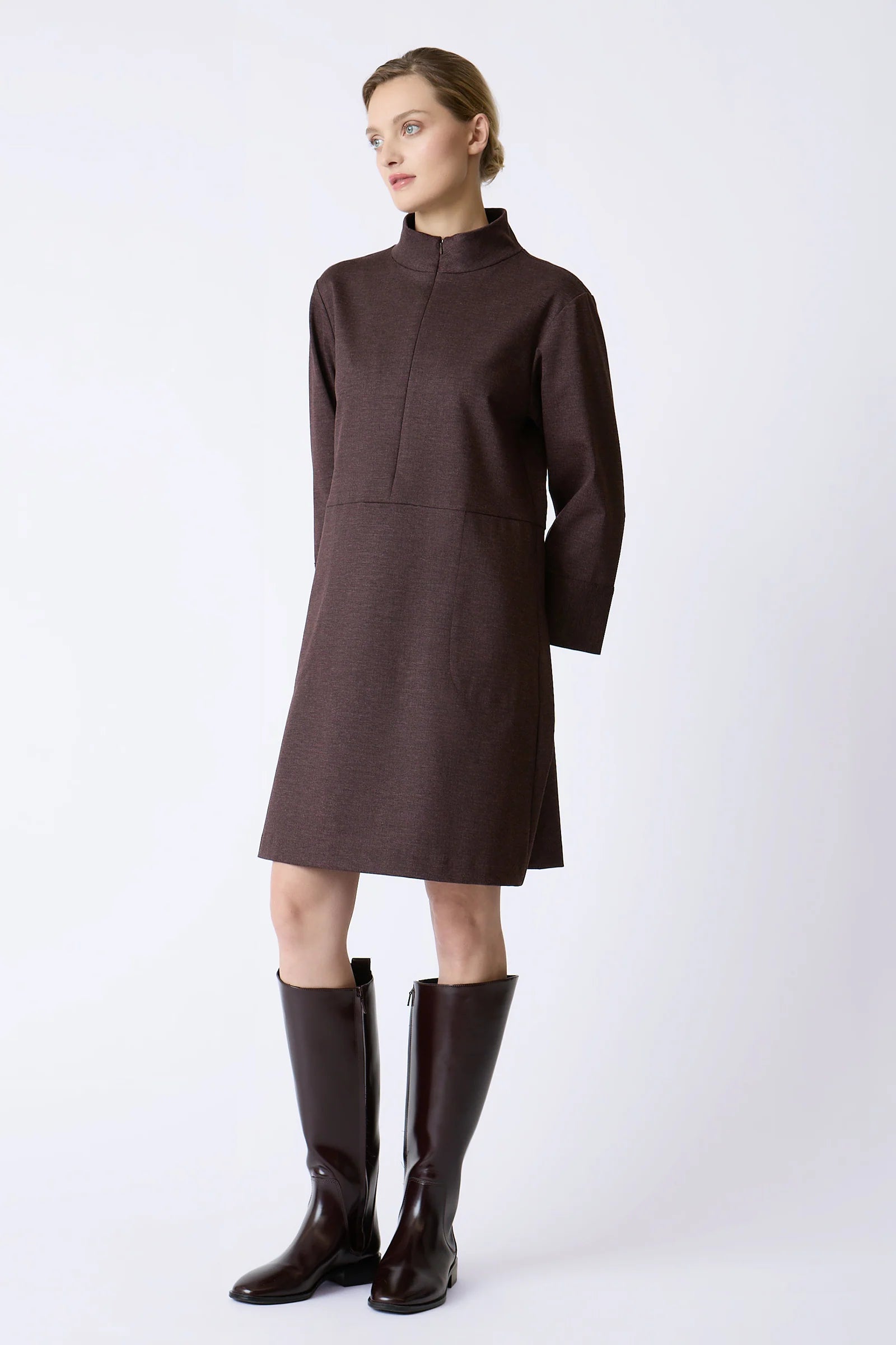 A woman modeling a burgundy high-neck, long-sleeved knee-length dress with a front zipper, paired with matching dark brown knee-high boots, against a plain light gray studio background.