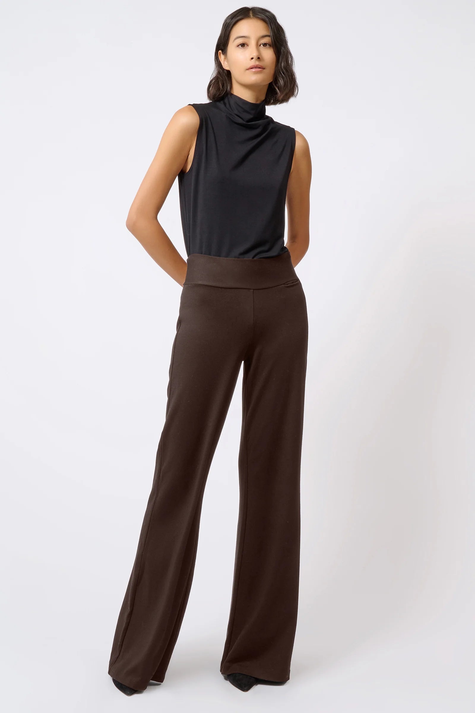 A model wearing a sleeveless black mock-neck top and dark brown wide-leg trousers, standing against a light gray studio background.