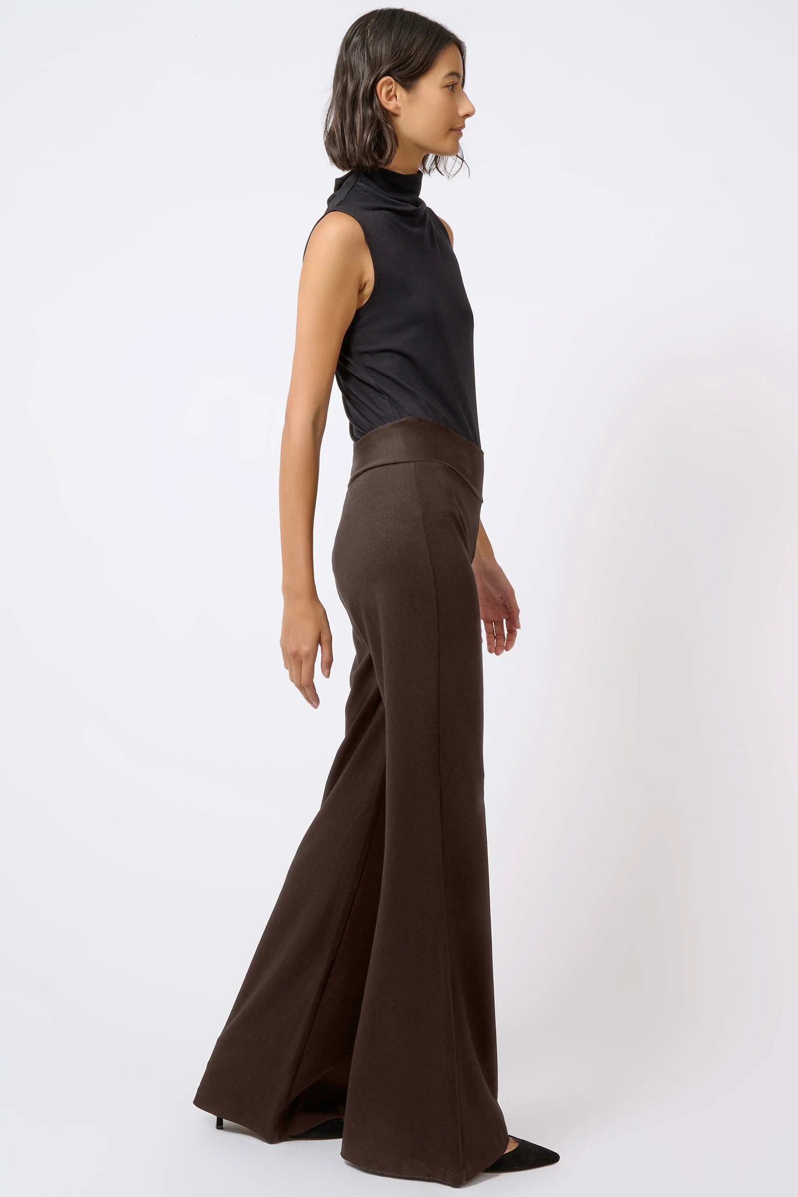 A model is shown from a side profile wearing a black sleeveless mock-neck top and dark brown wide-leg trousers against a plain white studio background.