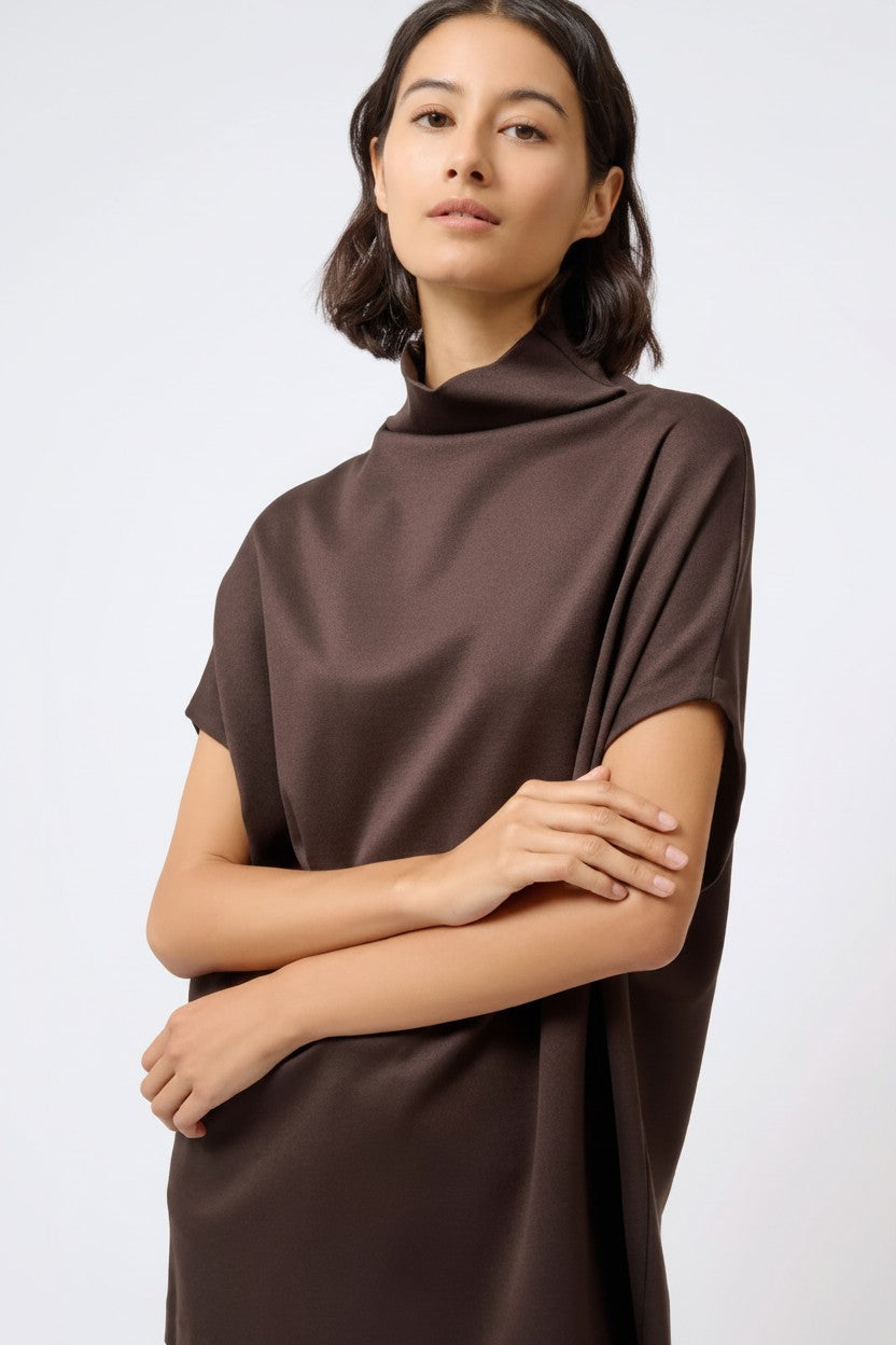 A woman wearing a dark brown short-sleeve mock neck dress or tunic top, posed against a light gray studio background.
