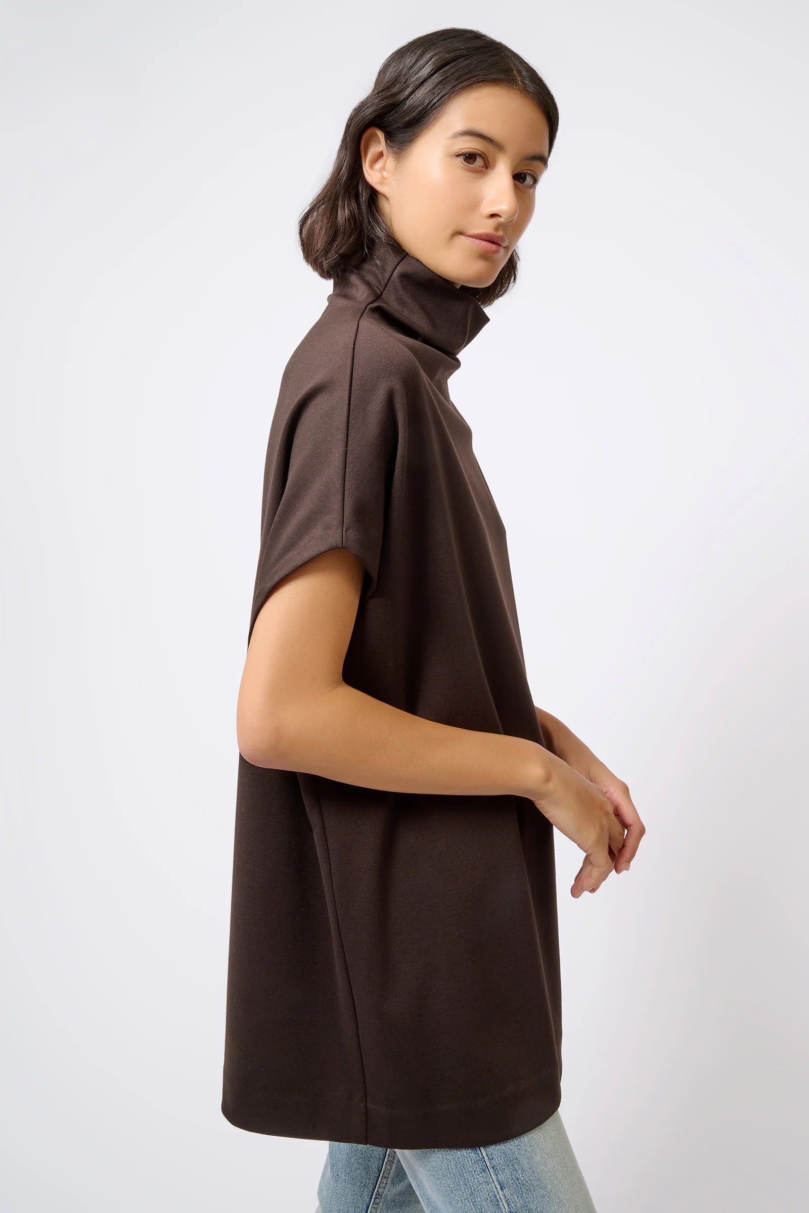 A woman wearing a dark brown short-sleeve mock neck tunic top, shown from a side profile against a light gray studio background.