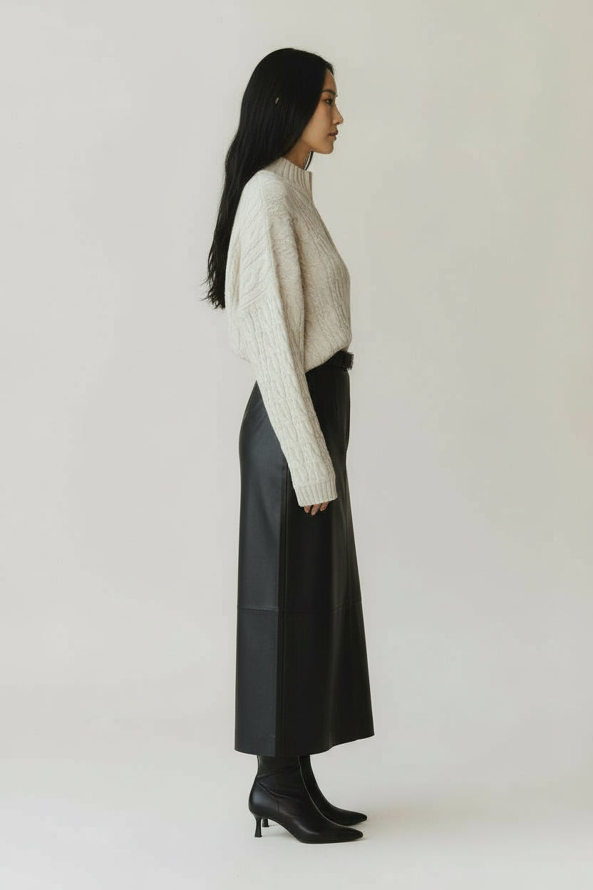 Woman wearing an ivory cable knit sweater with a maxi leather skirt and black boots on a gray background.