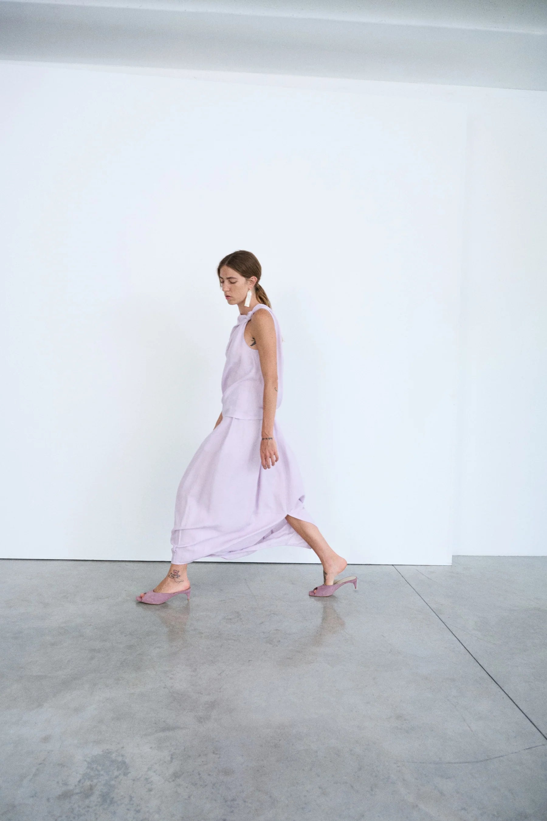 Woman in a light purple outfit walking on a reflective floor with a white wall background.