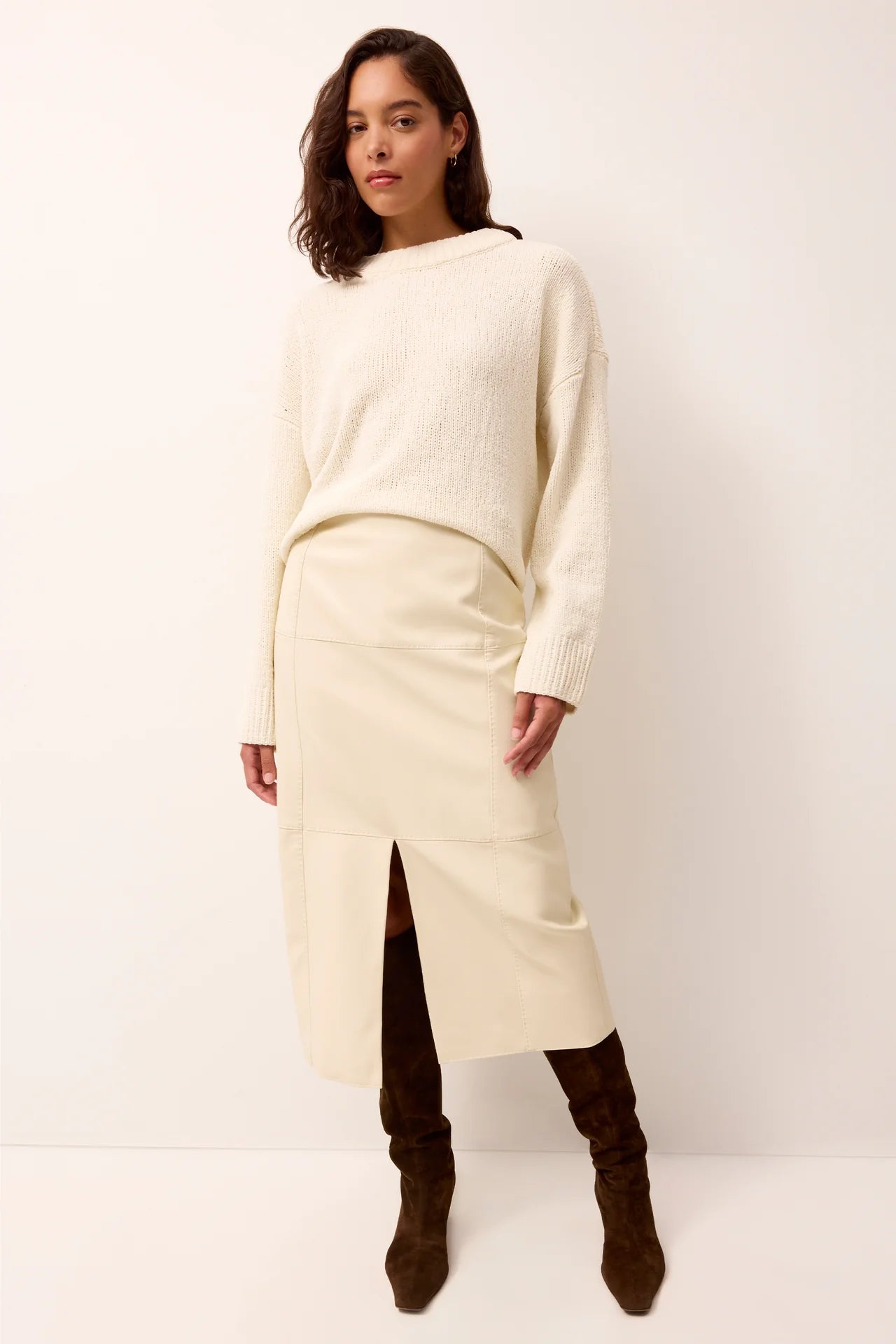 Woman wearing a cream sweater and skirt with brown boots on a white background.