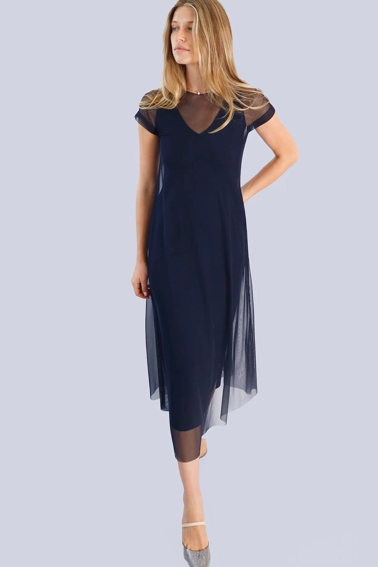 Woman wearing a navy blue dress with sheer sleeves on a light gray background.
