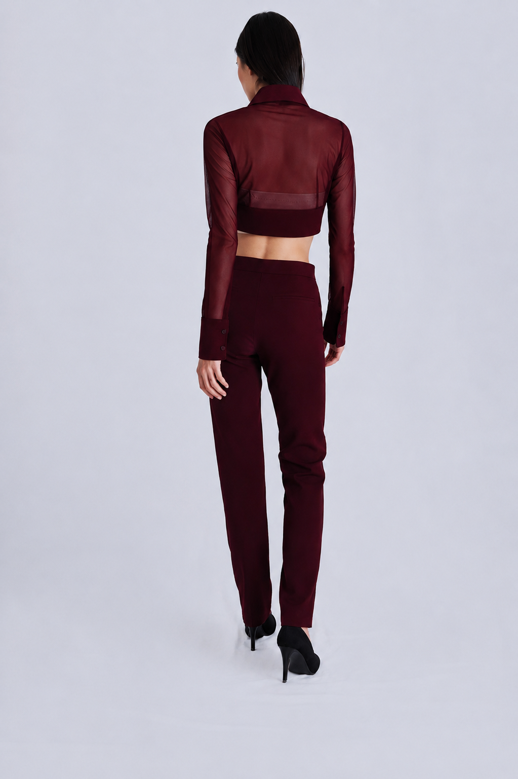 A model viewed from the back wearing a matching burgundy two-piece set consisting of a sheer long-sleeve crop top and high-waisted trousers, paired with black high heels on a light gray studio background.