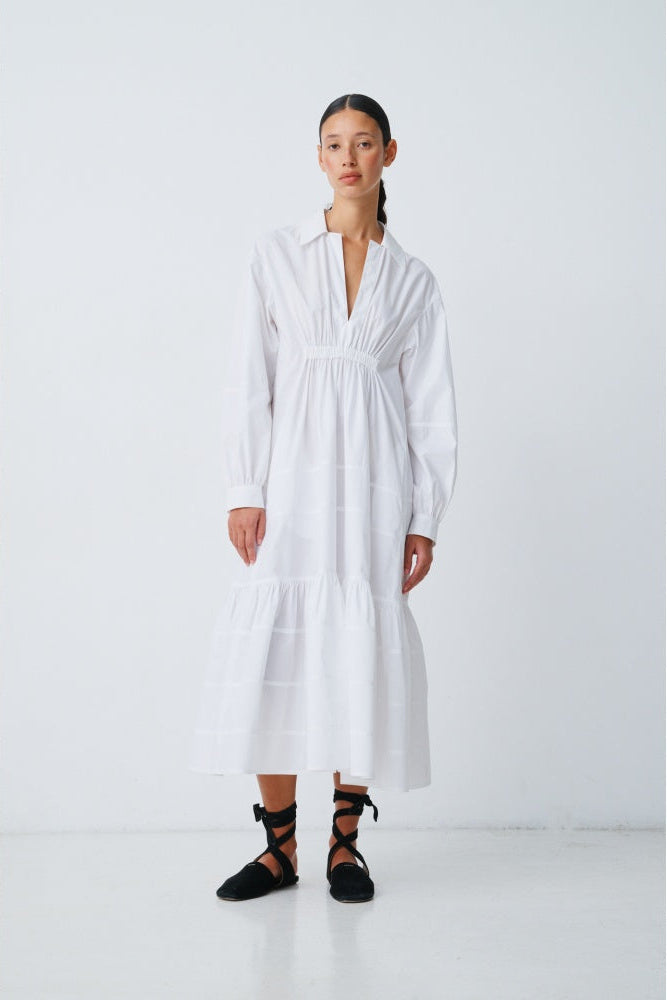 Woman wearing a white longsleeve shirt dress with black lace up shoes on a plain background.