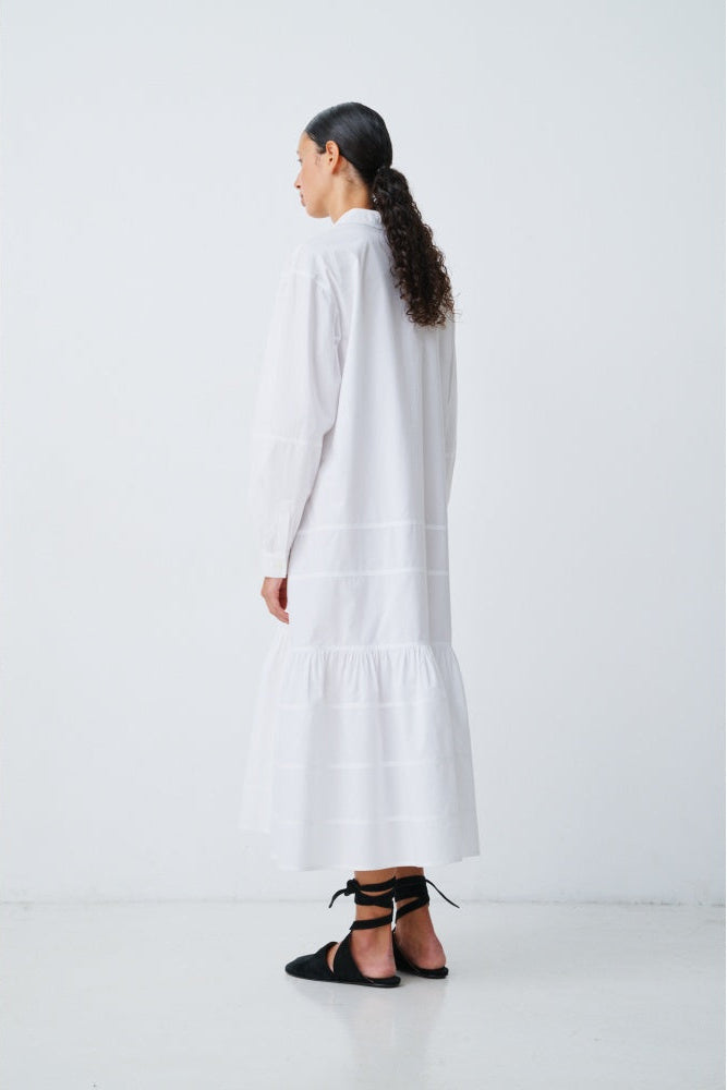The back of a woman wearing a white longsleeve dress with black shoes on a plain background.