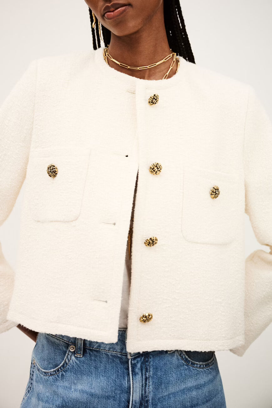 Woman wearing a white jacket with gold buttons and blue jeans on a white background.