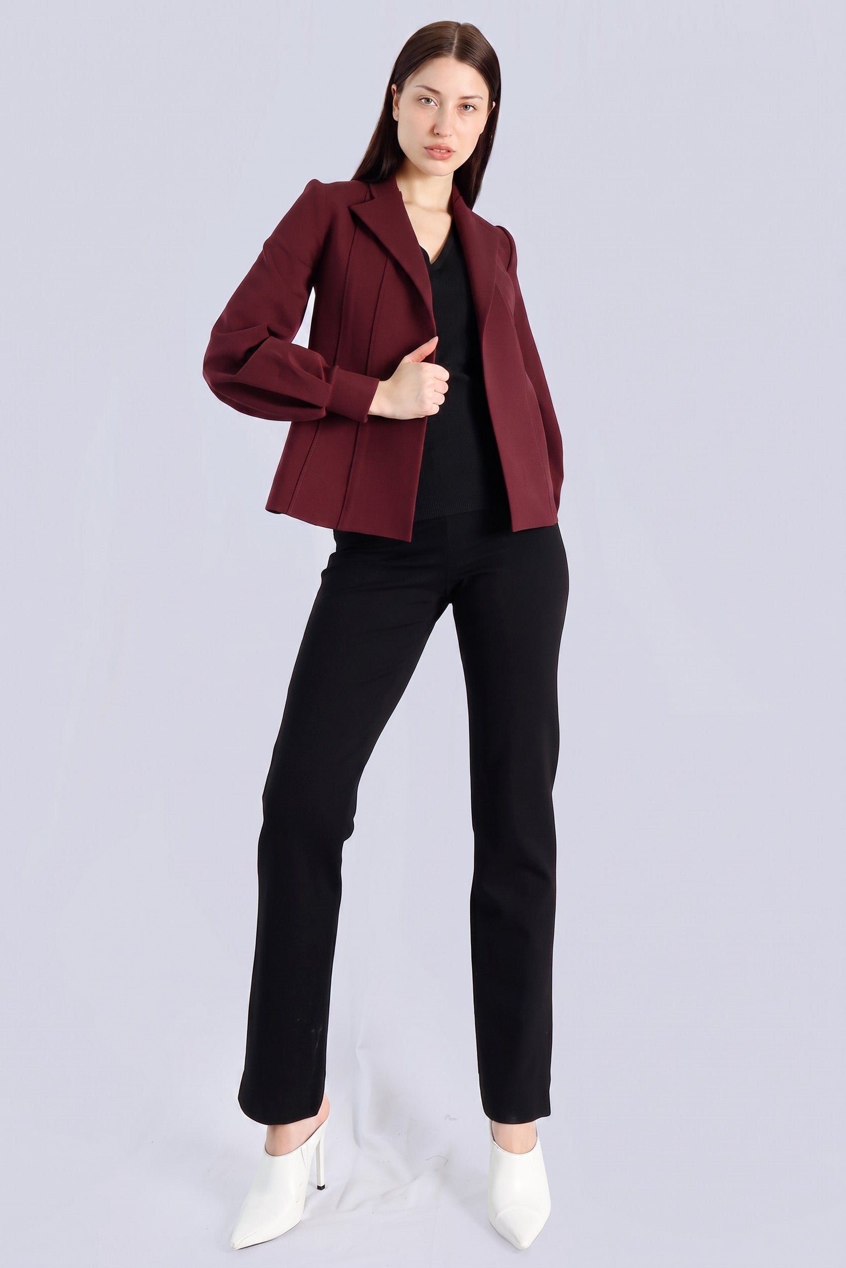 A woman modeling a structured, cropped maroon blazer over a black top and black trousers, paired with white pointed-toe heels, against a light gray studio background.