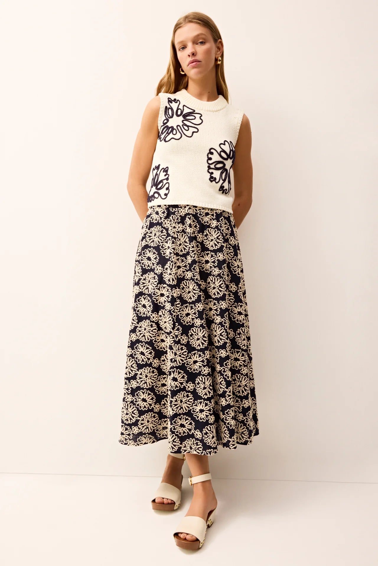 Woman wearing a white sleeveless sweater with navy floral design and a matching navy and white patterned skirt on a white background.