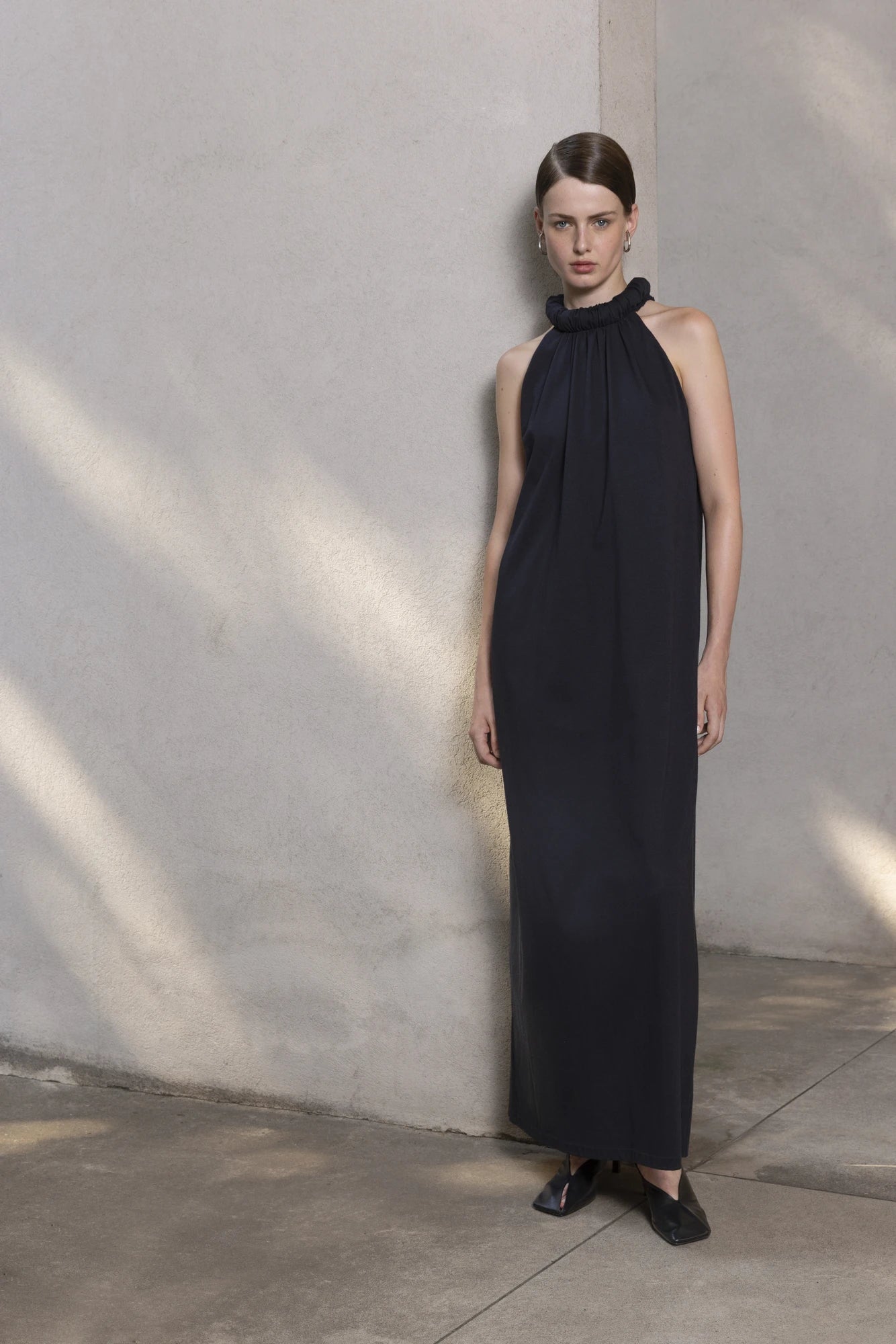 Woman wearing a black full length dress with black flats against a stone wall.